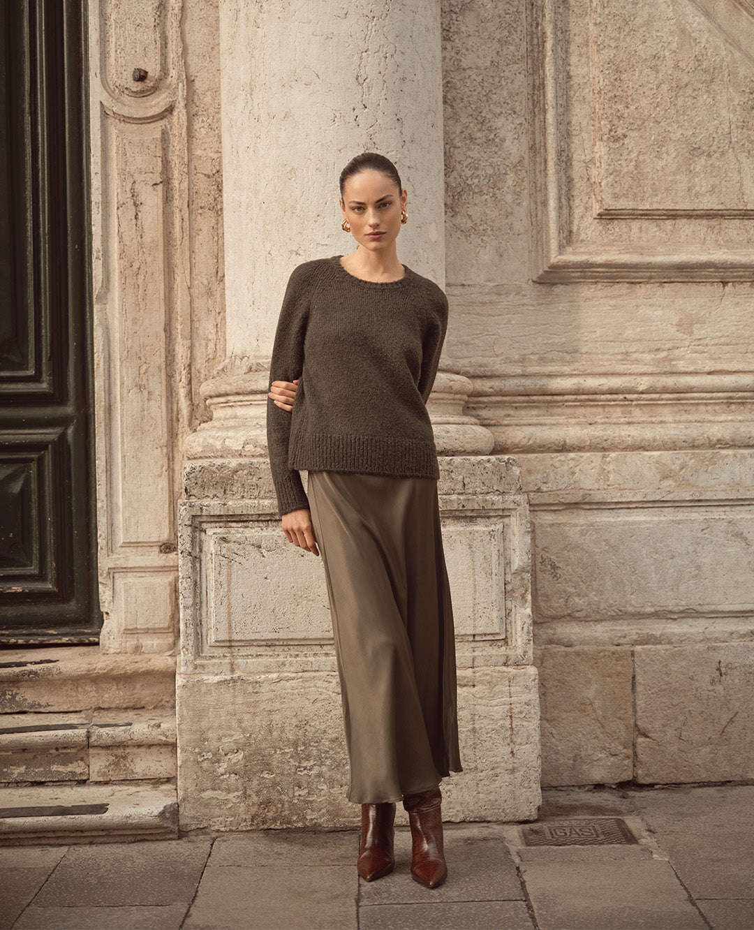 Eine Frau steht vor einer hellen Steinfassade und trägt einen olivgrünen Pullover mit fließendem Rock und braunen Stiefeletten. Der Look ist elegant, schlicht und herbstlich modern.