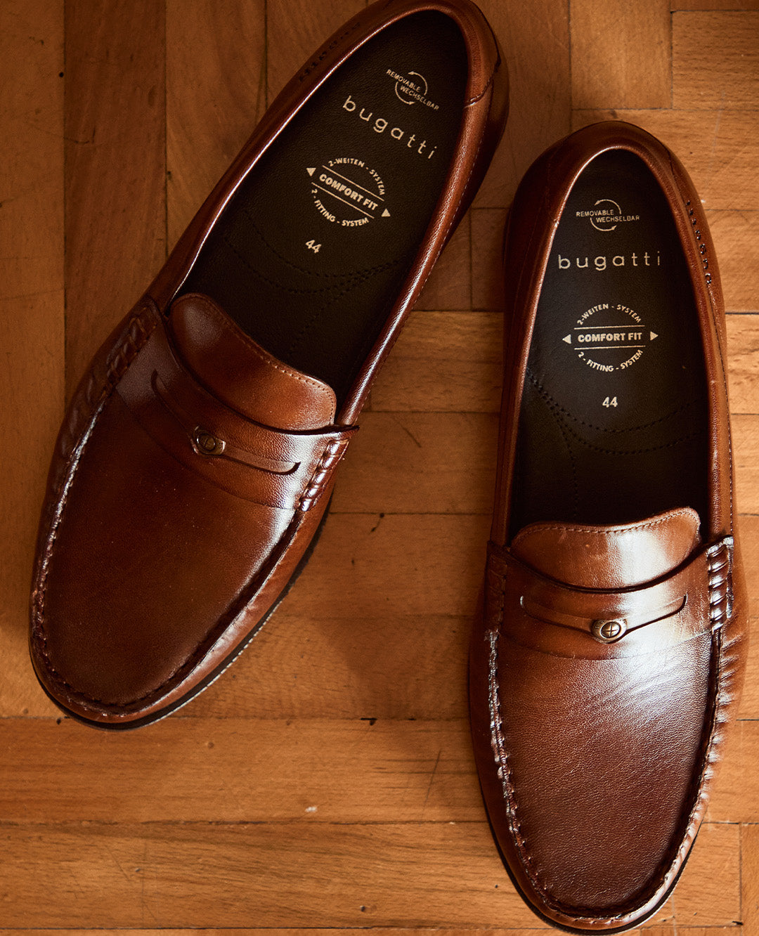 Ein Paar braune bugatti Loafer aus Leder liegt auf einem Holzboden. Die Schuhe strahlen klassische Eleganz, hochwertige Verarbeitung und zeitlosen Stil aus.