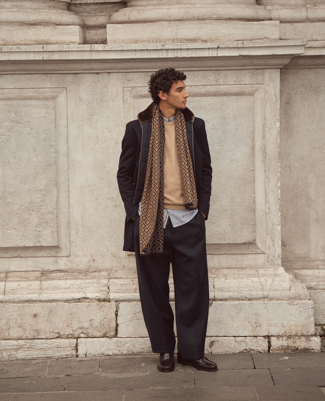 Ein Mann trägt eine dunkelblaue Stoffhose, einen hellbraunen Pullover und einen gemusterten Schal über einem gestreiften Hemd. Der Look kombiniert klassische Eleganz mit moderner Lässigkeit – perfekt für den Herbst in der Stadt.