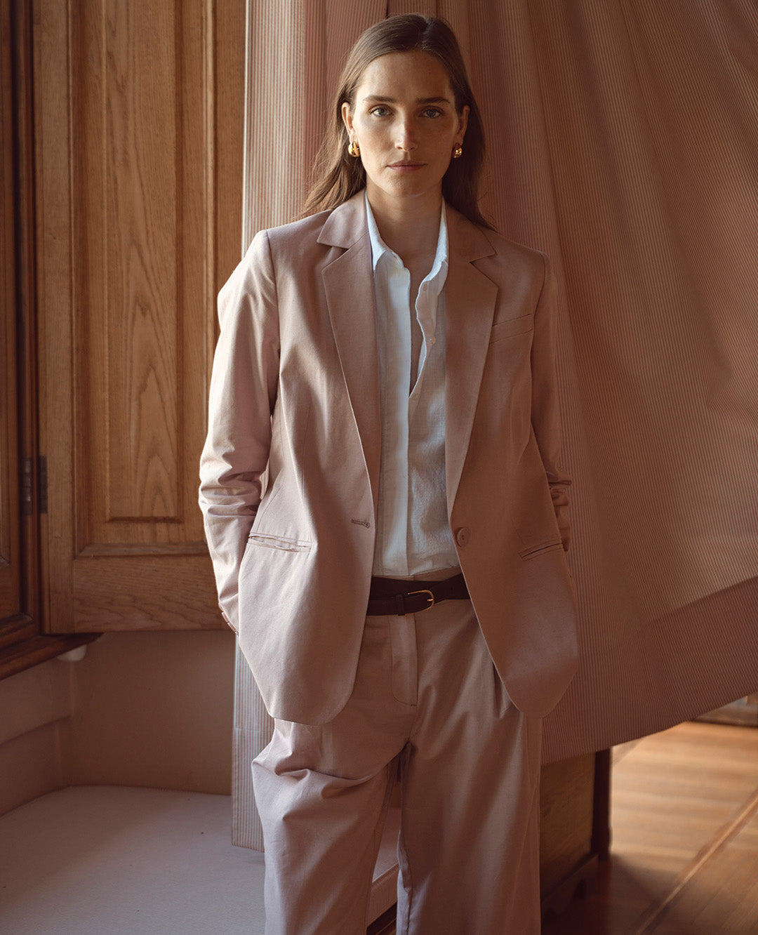 Eine Frau trägt einen hellrosa Hosenanzug mit weißer Bluse und dunklem Gürtel. Der Look wirkt elegant, modern und zugleich dezent feminin.