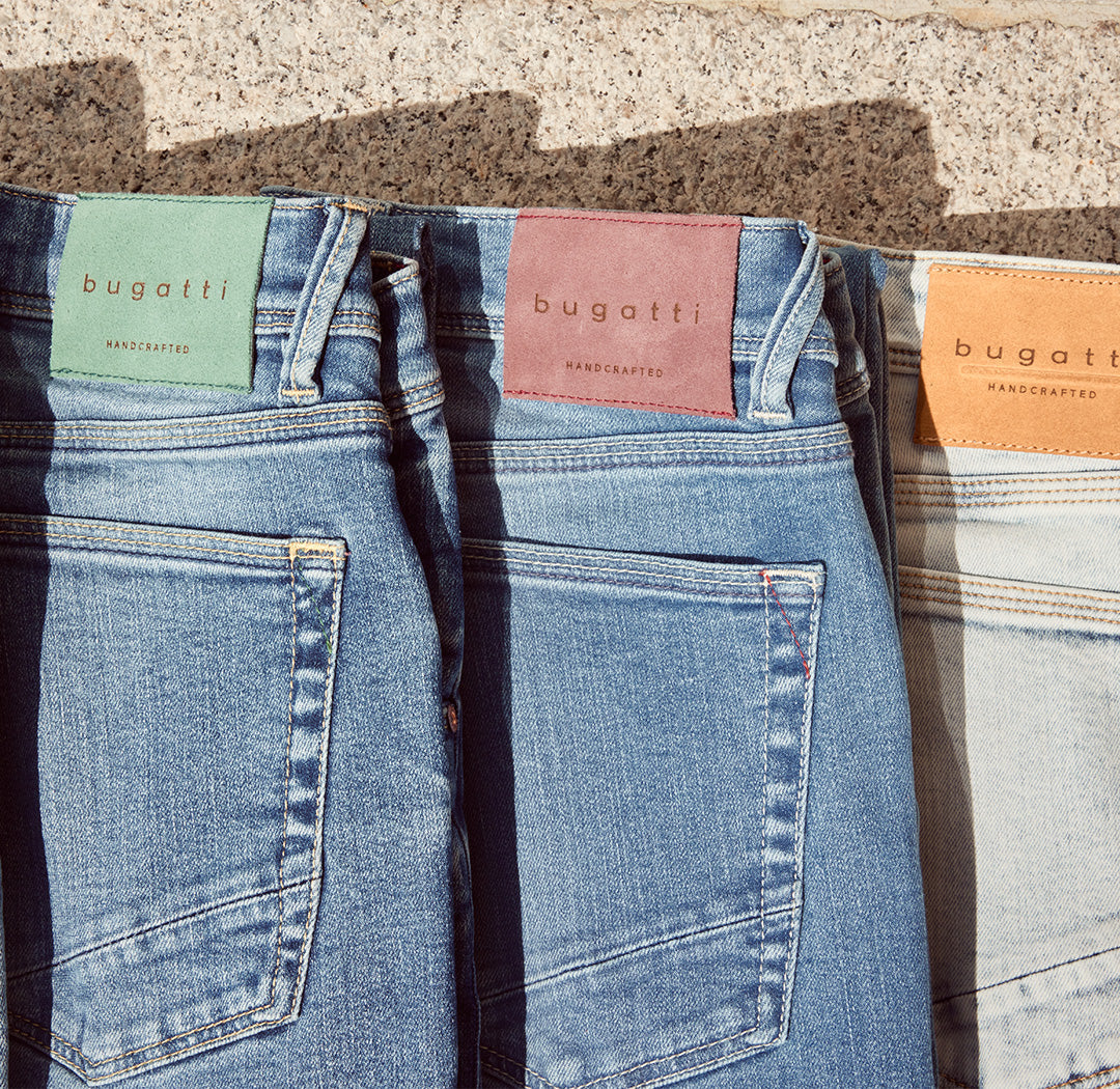 Drei bugatti Jeans liegen nebeneinander, jede mit farbigem Label in Grün, Rot und Orange. Das Bild betont Handwerkskunst, Qualität und sommerliche Lässigkeit.