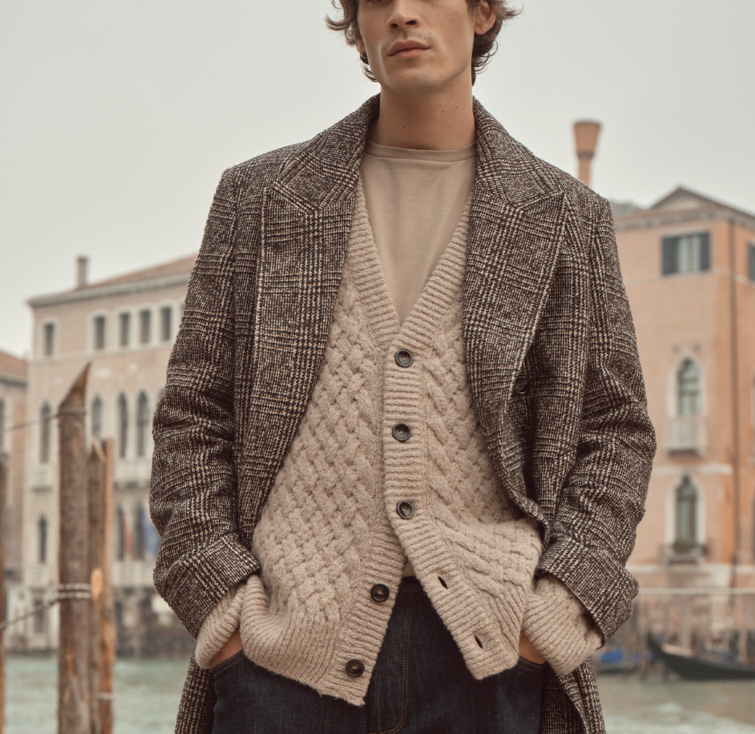 Ein Mann trägt einen karierten Mantel über einer beigen Strickjacke und einem hellen Shirt. Der Look kombiniert klassische Eleganz mit moderner Lässigkeit und herbstlichem Charme.