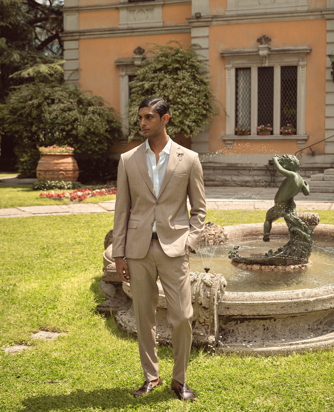 Ein Mann steht vor einem Brunnen im Garten und trägt einen hellbeigen Anzug mit weißem Hemd. Der Look ist elegant, sommerlich und klassisch stilvoll.