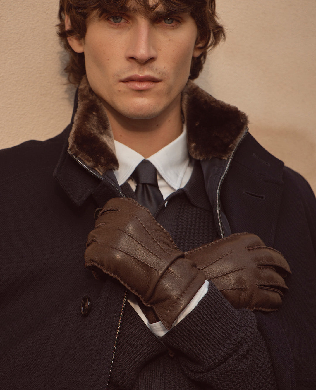 Nahaufnahme eines Mannes in dunklem Mantel mit Fellkragen, weißem Hemd und Krawatte. Er trägt braune Lederhandschuhe – ein eleganter, markanter Winterlook.