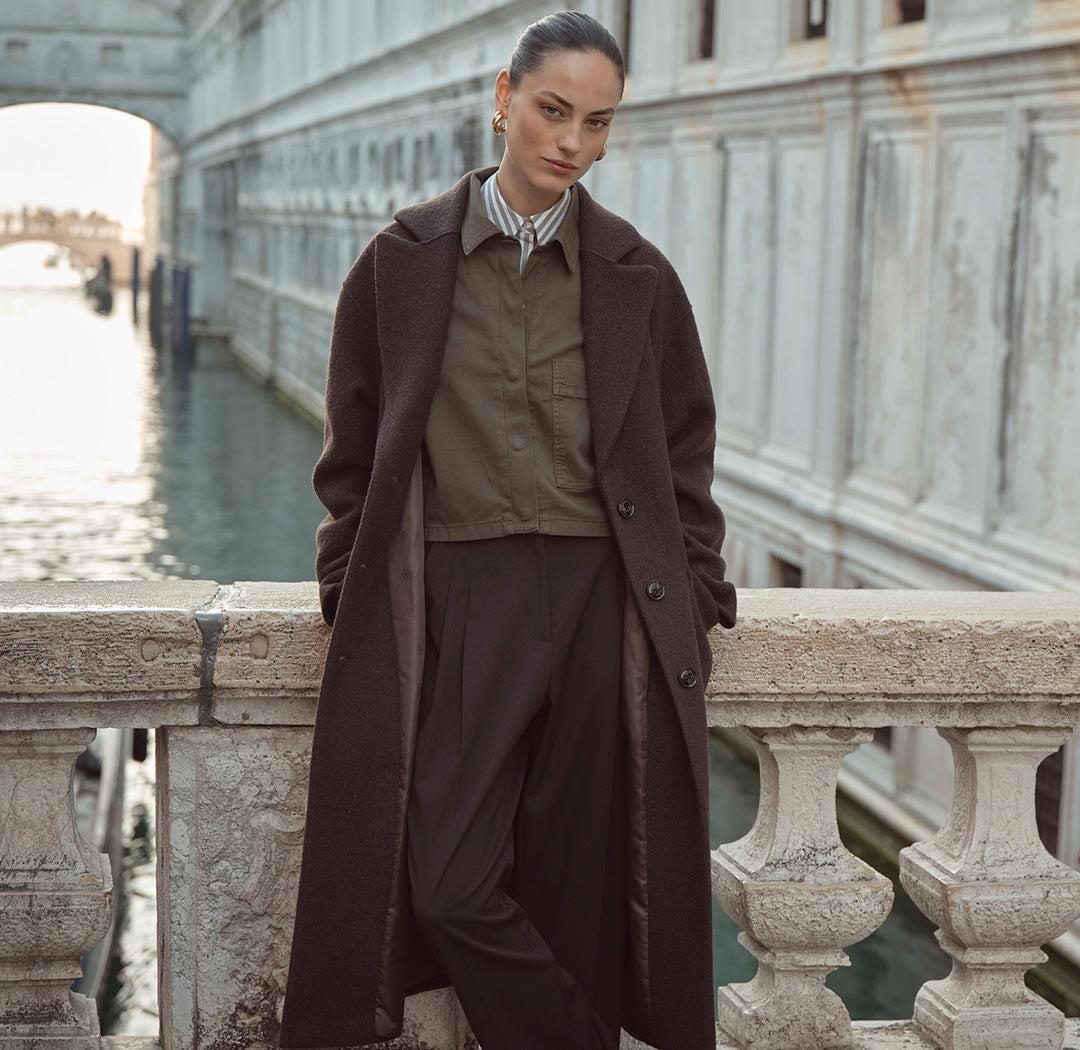 Eine Frau steht auf einer Brücke vor historischer Kulisse und trägt einen langen dunklen Mantel über einem olivgrünen Hemd und dunkler Hose. Der Look ist elegant und urban.