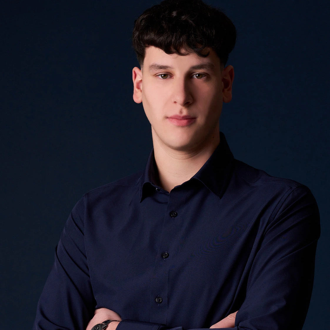 Young man with short dark hair wearing a navy button-up shirt and watch against a dark backdrop