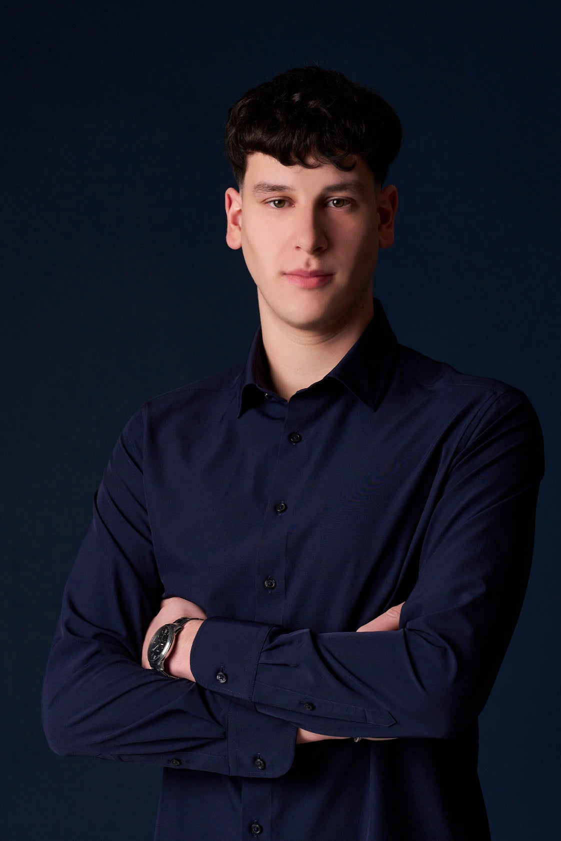 Young man with short dark hair wearing a navy button-up shirt and watch standing against a dark background