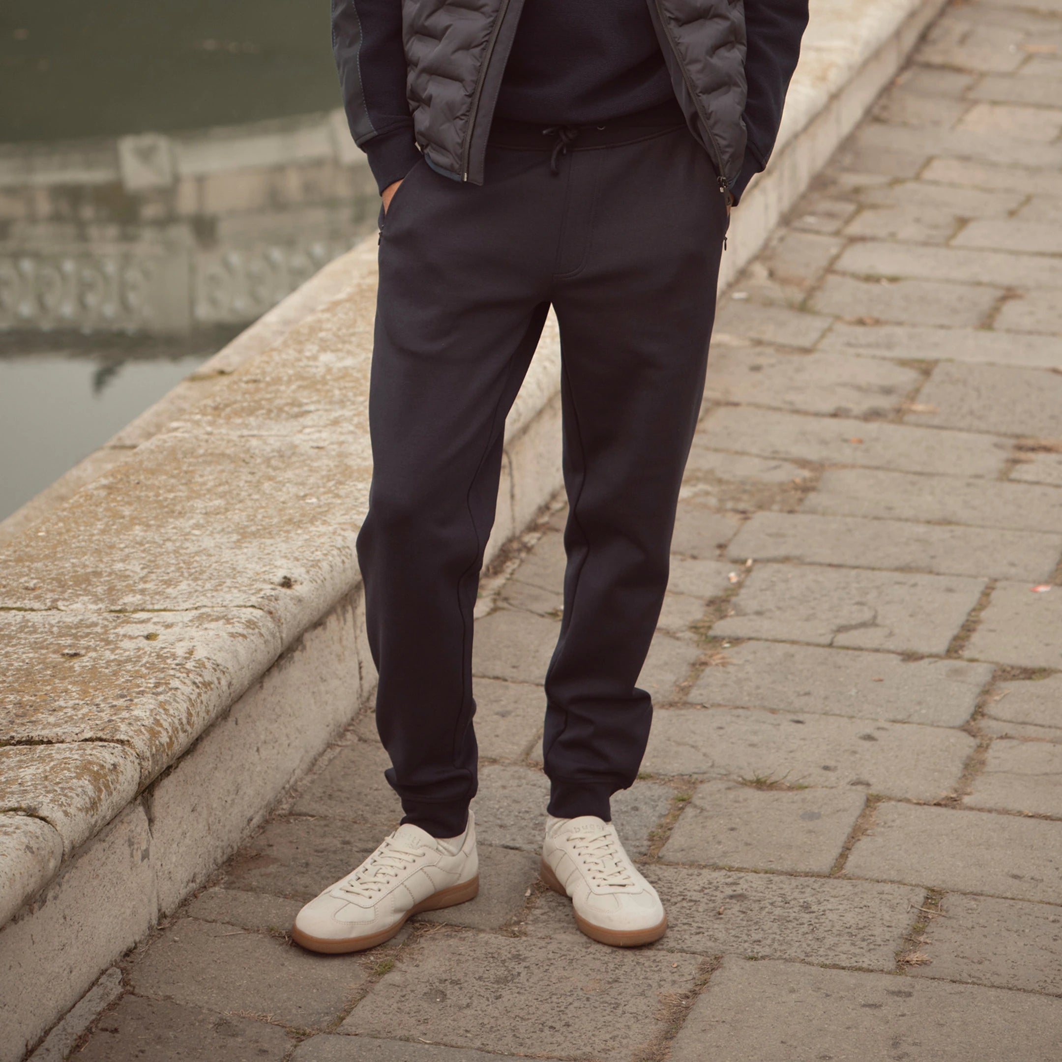 Mann trägt dunkelblaue bugatti Jogpants kombiniert mit weißen Sneakers und gesteppter Weste – moderner Casual Look der Herren Herbst/Winter-Kollektion 2025 im Venetian Chic.
