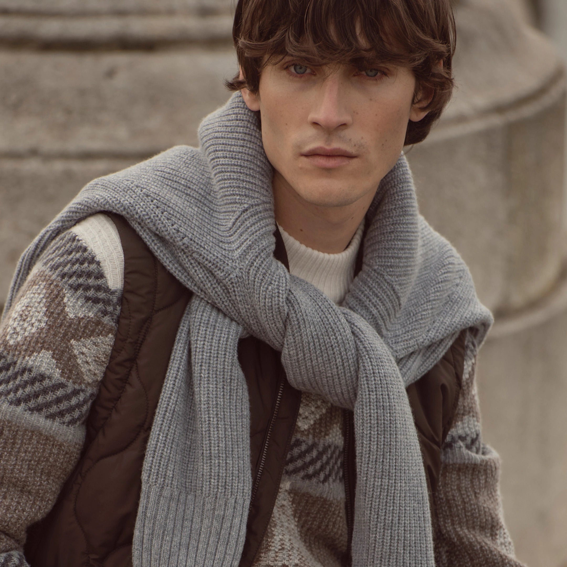 Mann trägt bugatti Strickpullover mit grafischem Muster in Naturtönen, darüber gesteppte Weste und über die Schultern gelegten grauen Schal – stylischer Herrenlook der Herbst/Winter-Kollektion 2025 im Venetian Chic.