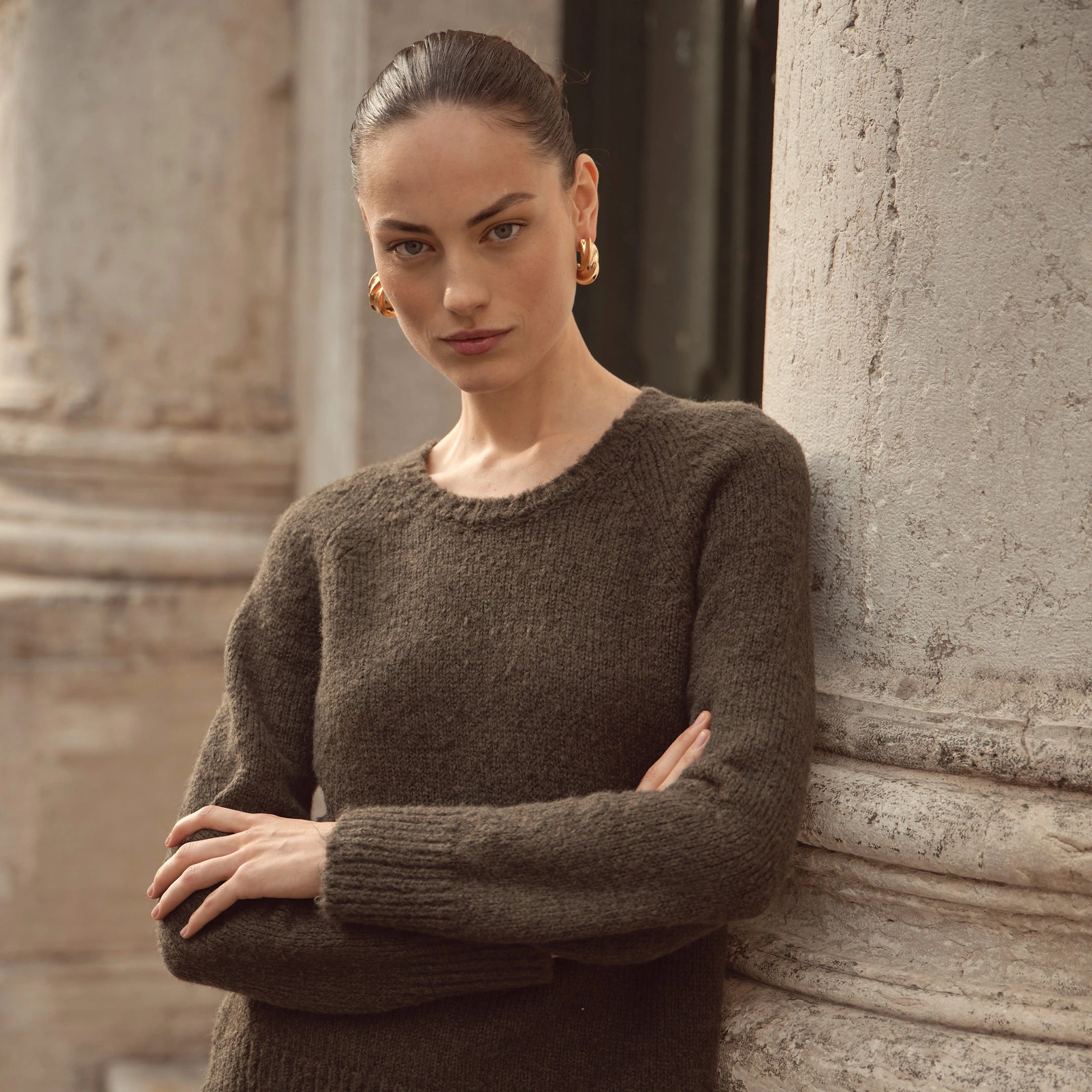 Frau trägt dunkelolivgrünen bugatti Strickpullover mit Rundhalsausschnitt, kombiniert mit goldenen Creolen – eleganter Look der Damen Herbst/Winter-Kollektion 2025 im Venetian Chic.