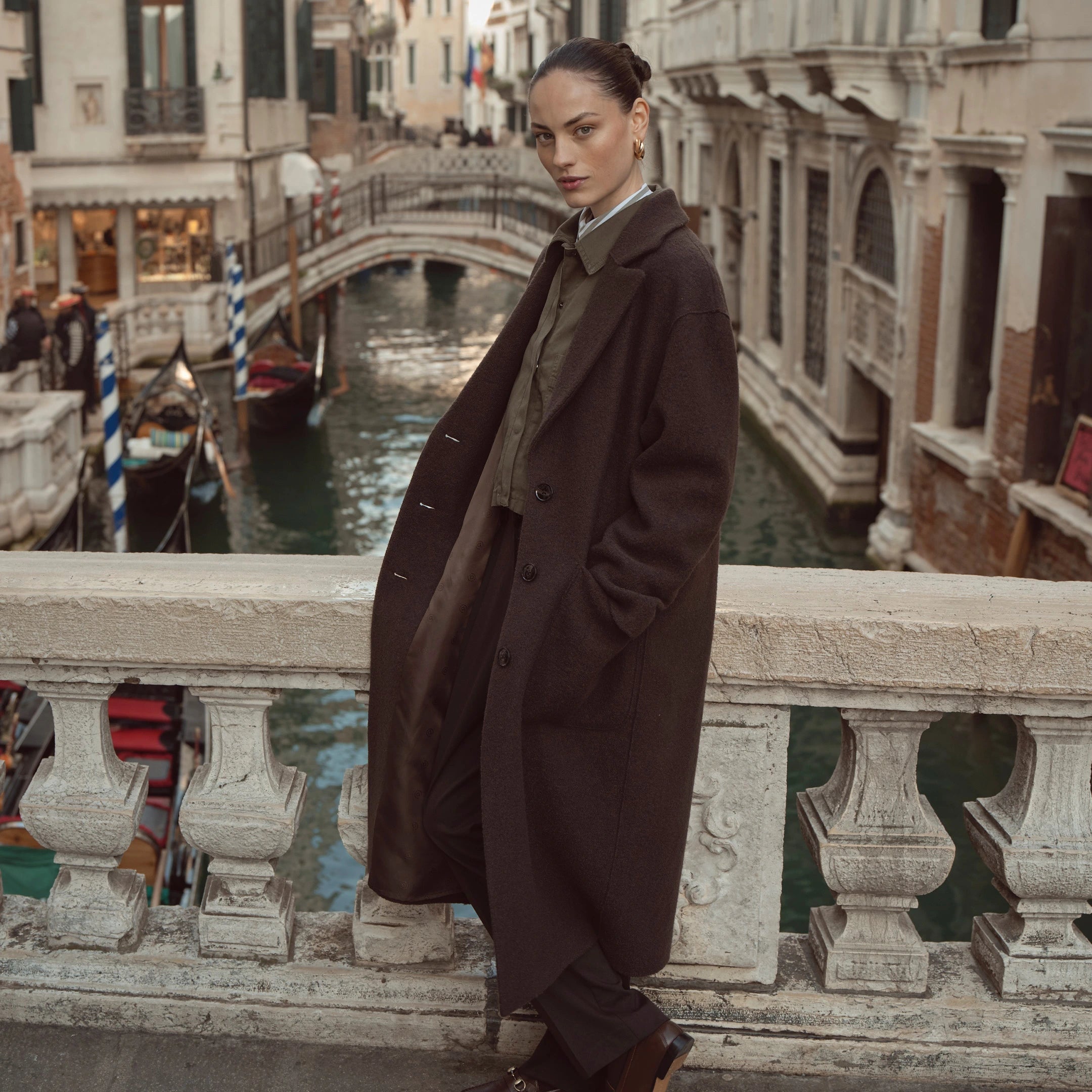 Frau trägt langen dunkelbraunen bugatti Mantel über olivfarbener Bluse und dunkler Hose, aufgenommen auf einer Brücke in Venedig – eleganter Damenlook der Herbst/Winter-Kollektion 2025 im Venetian Chic.