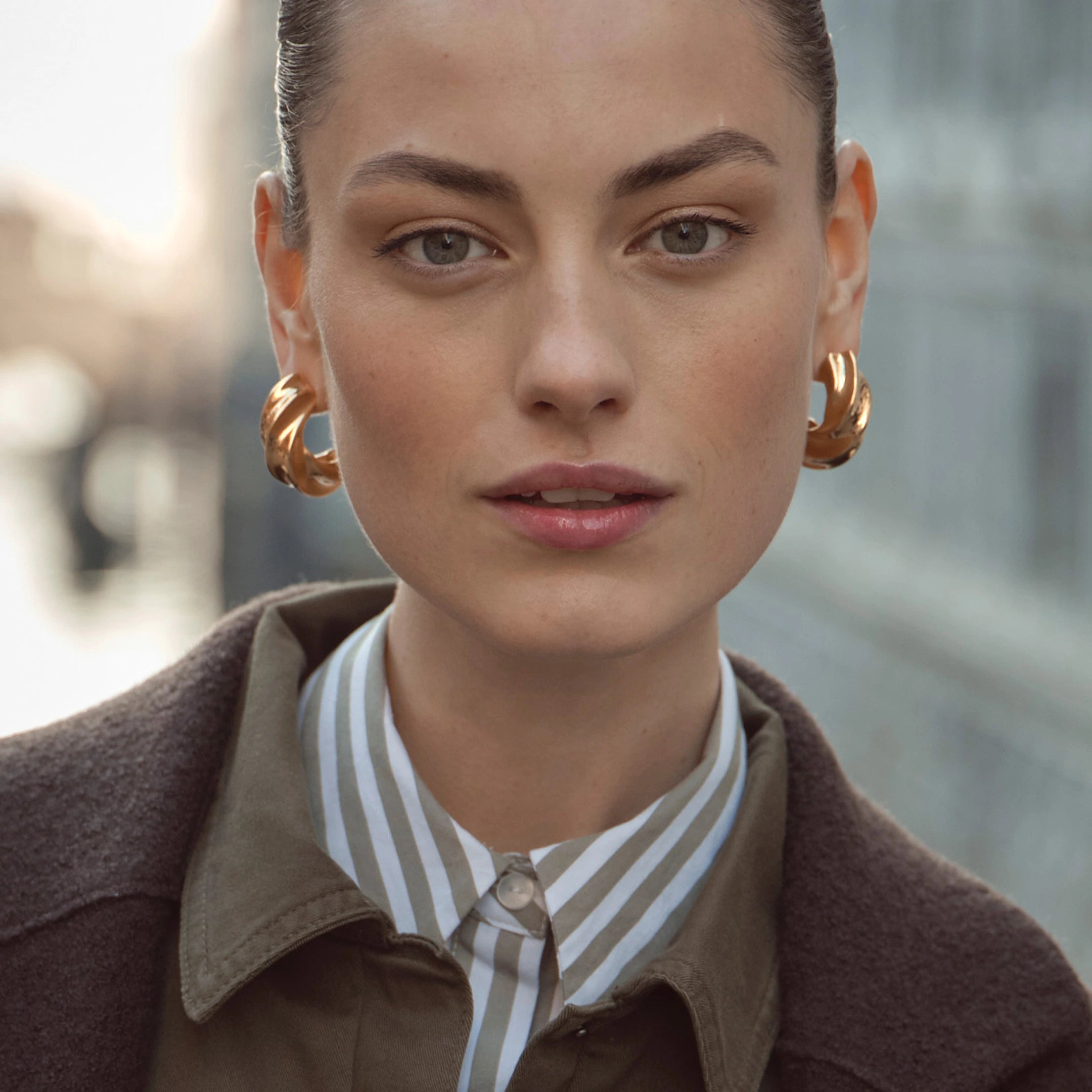 Nahaufnahme einer Frau mit goldfarbenen Creolen, gestreifter Bluse in Grau-Weiß und olivgrünem Blazer unter dunkelbraunem Mantel – moderner bugatti Look der Herbst/Winter-Kollektion 2025 im venezianischen Setting.