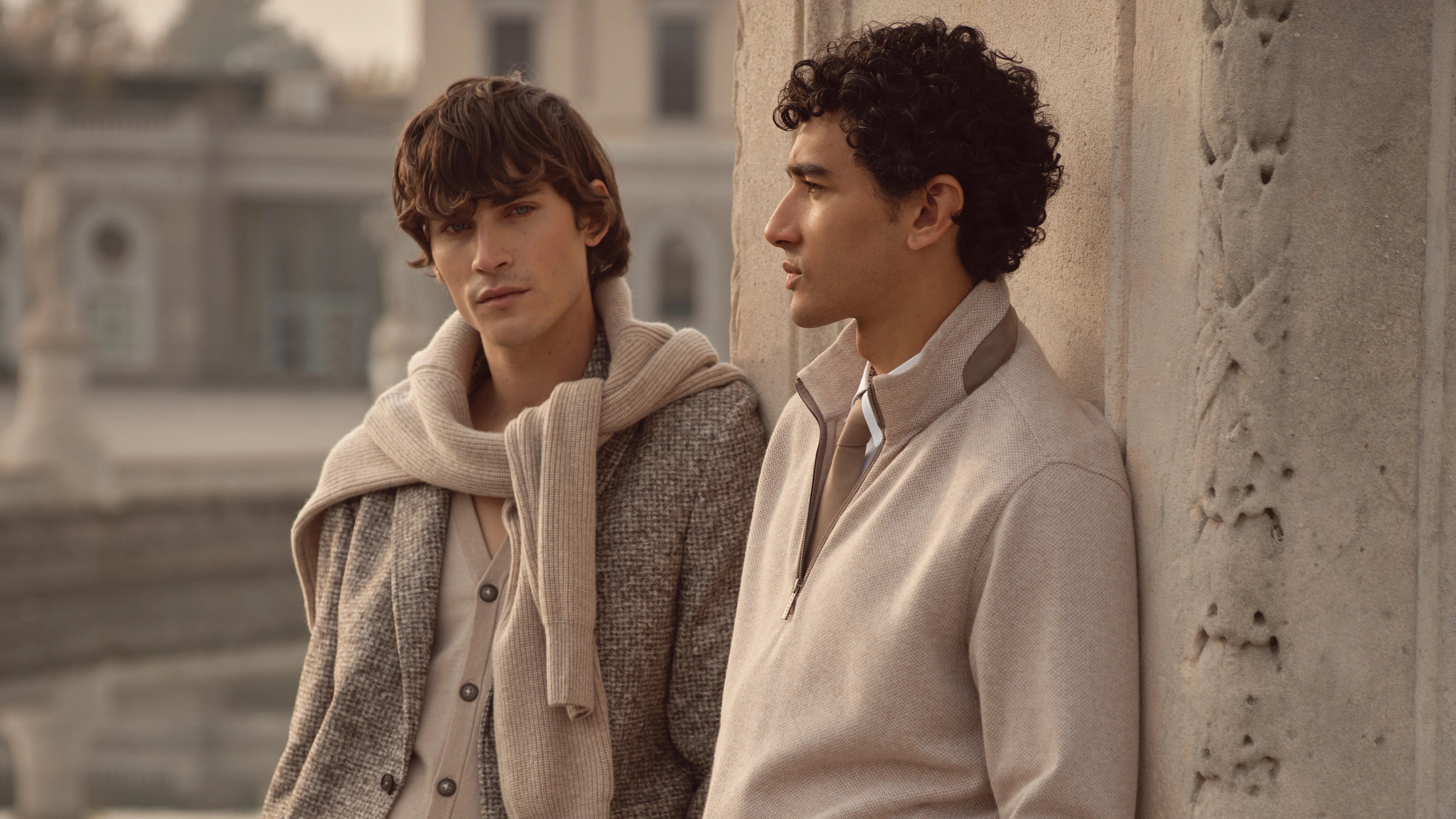 Zwei Männer tragen bugatti Herbst/Winter-Looks 2025: links Sakko mit Strickpullover über die Schultern, rechts beiger Zipper-Pullover mit Hemd und Krawatte – elegante Menswear im Venetian Chic.