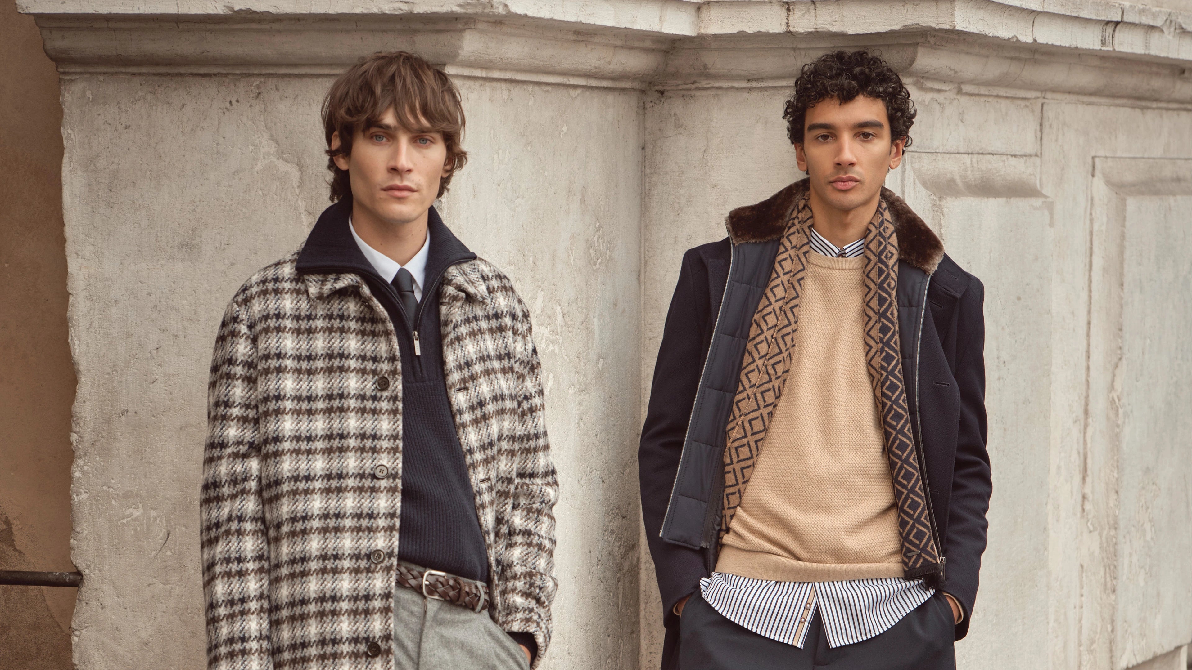 Zwei Männer tragen bugatti Herbst/Winter-Looks 2025: links karierten Mantel über Hemd, Pullover und Krawatte, rechts dunkelblauen Mantel mit Fellkragen, beigem Pullover und gemustertem Schal – elegante Menswear im Venetian Chic.