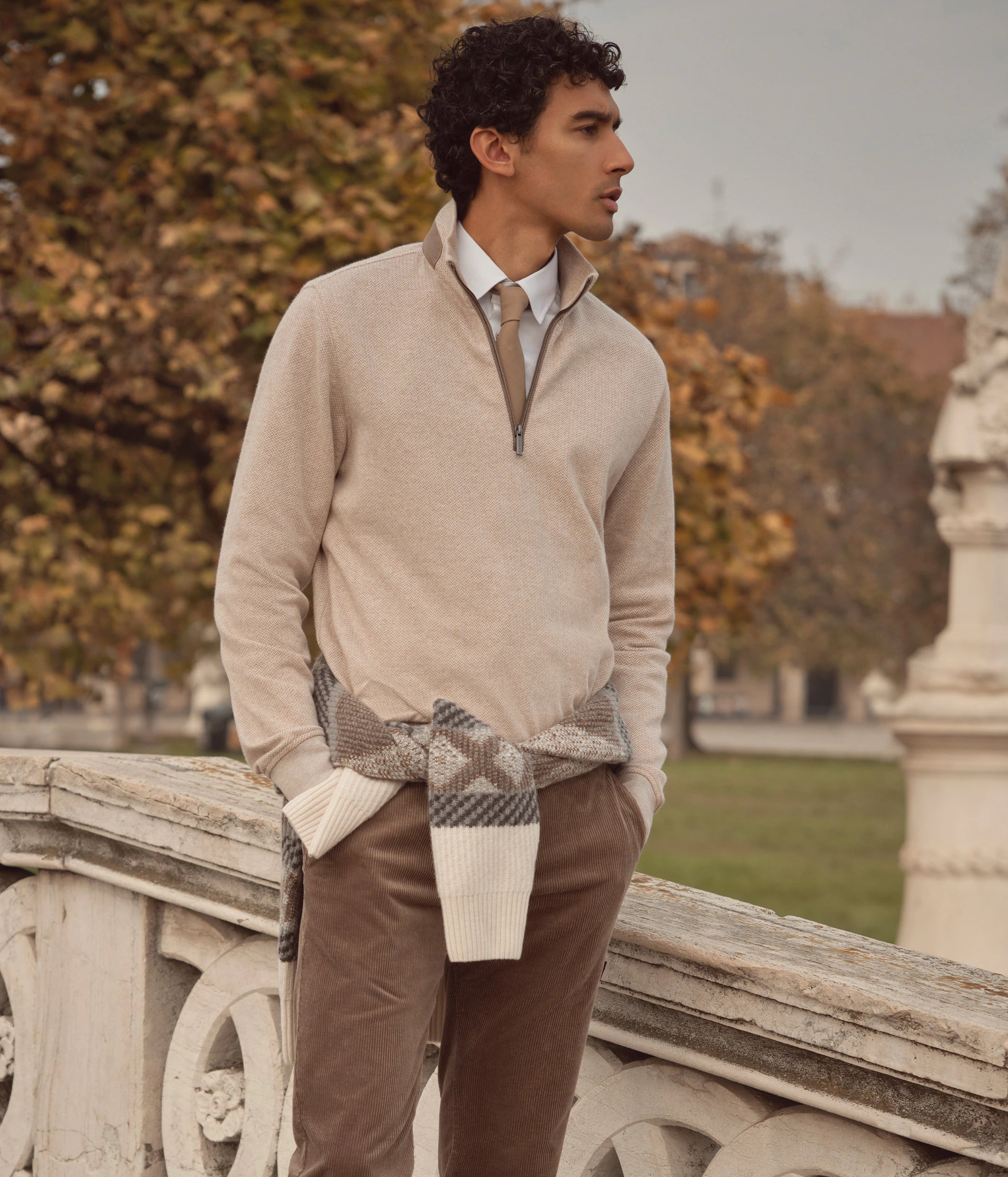 Mann trägt beigefarbenen bugatti-Halbzip-Pullover über weißem Hemd mit Krawatte, kombiniert mit brauner Cordhose – herbstlicher Look mit Strick-Details, aufgenommen im Freien vor herbstlicher Kulisse.