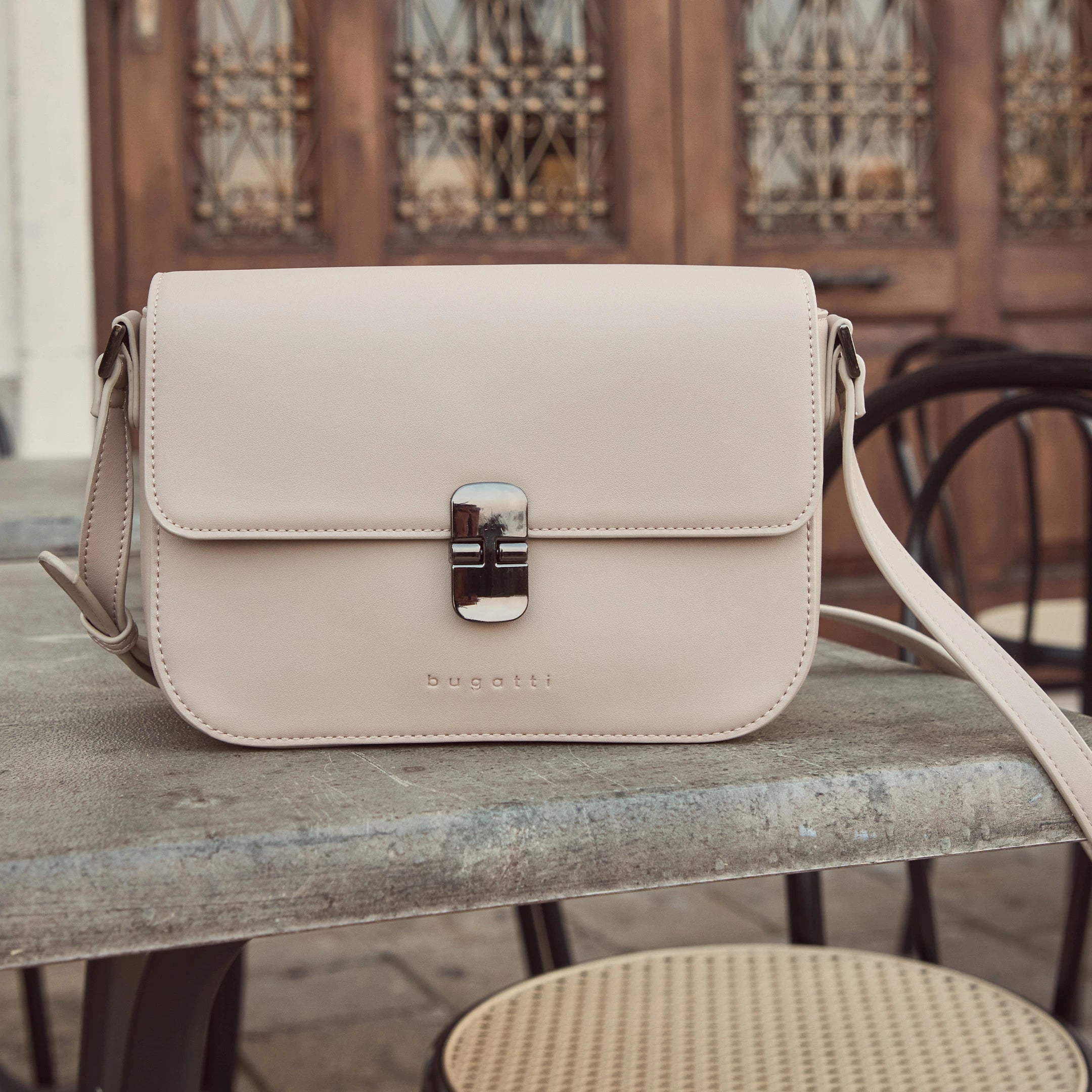 Beige bugatti Umhängetasche mit silberner Metallschließe, elegant auf einem Café-Tisch inszeniert – stilvolles Accessoire der Damen Herbst/Winter-Kollektion 2025 im Venetian Chic.