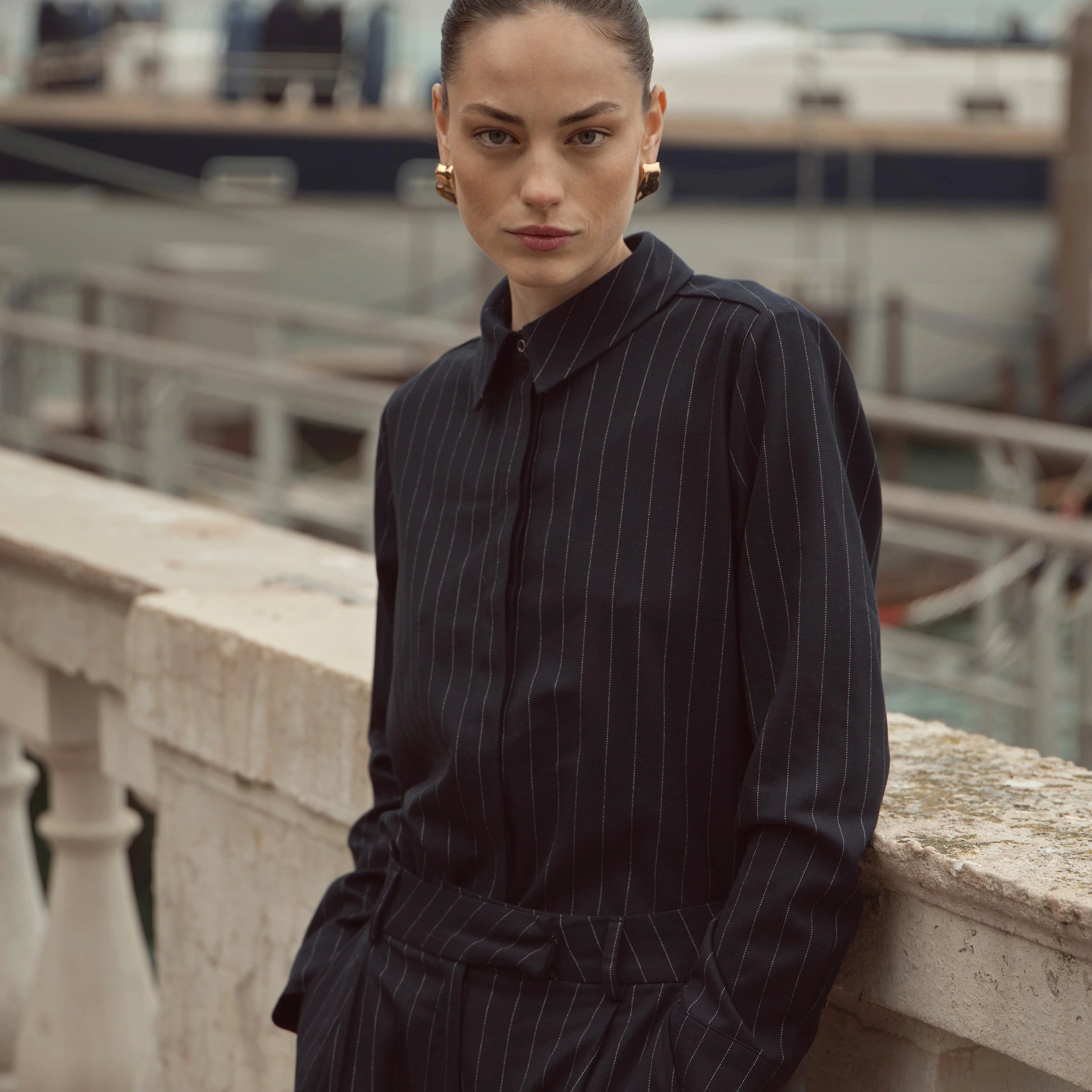 Frau trägt navyfarbene bugatti Bluse mit feinen Nadelstreifen aus der Damen Herbst/Winter-Kollektion 2025 – eleganter Venetian Chic Look, stilvoll kombiniert mit goldenen Ohrringen.