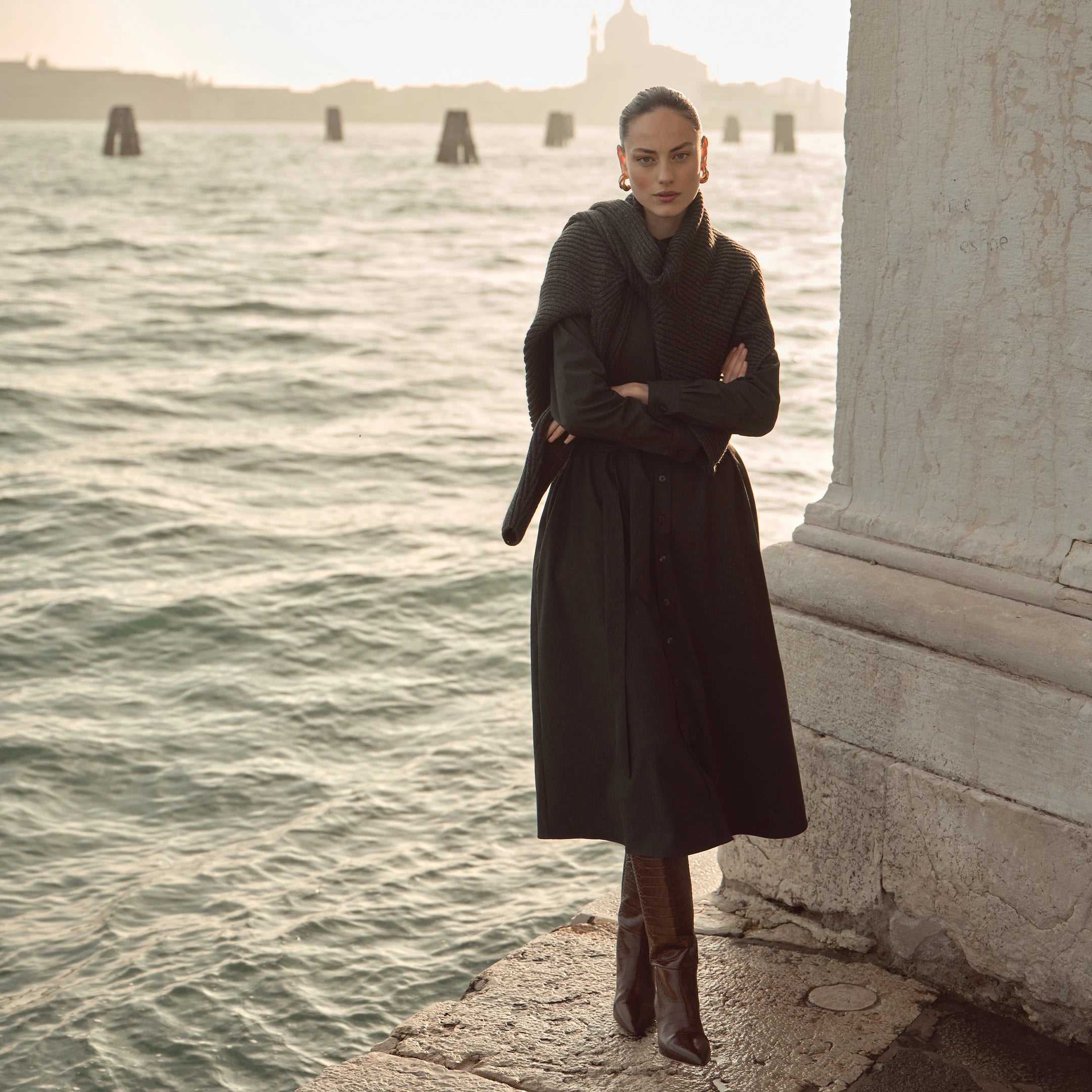 Frau trägt schwarzes bugatti Hemdblusenkleid in Midilänge mit über die Schultern gelegtem Strickpullover, kombiniert mit braunen Stiefeln – eleganter Look der Damen Herbst/Winter-Kollektion 2025 im Venetian Chic am Wasser in Venedig.