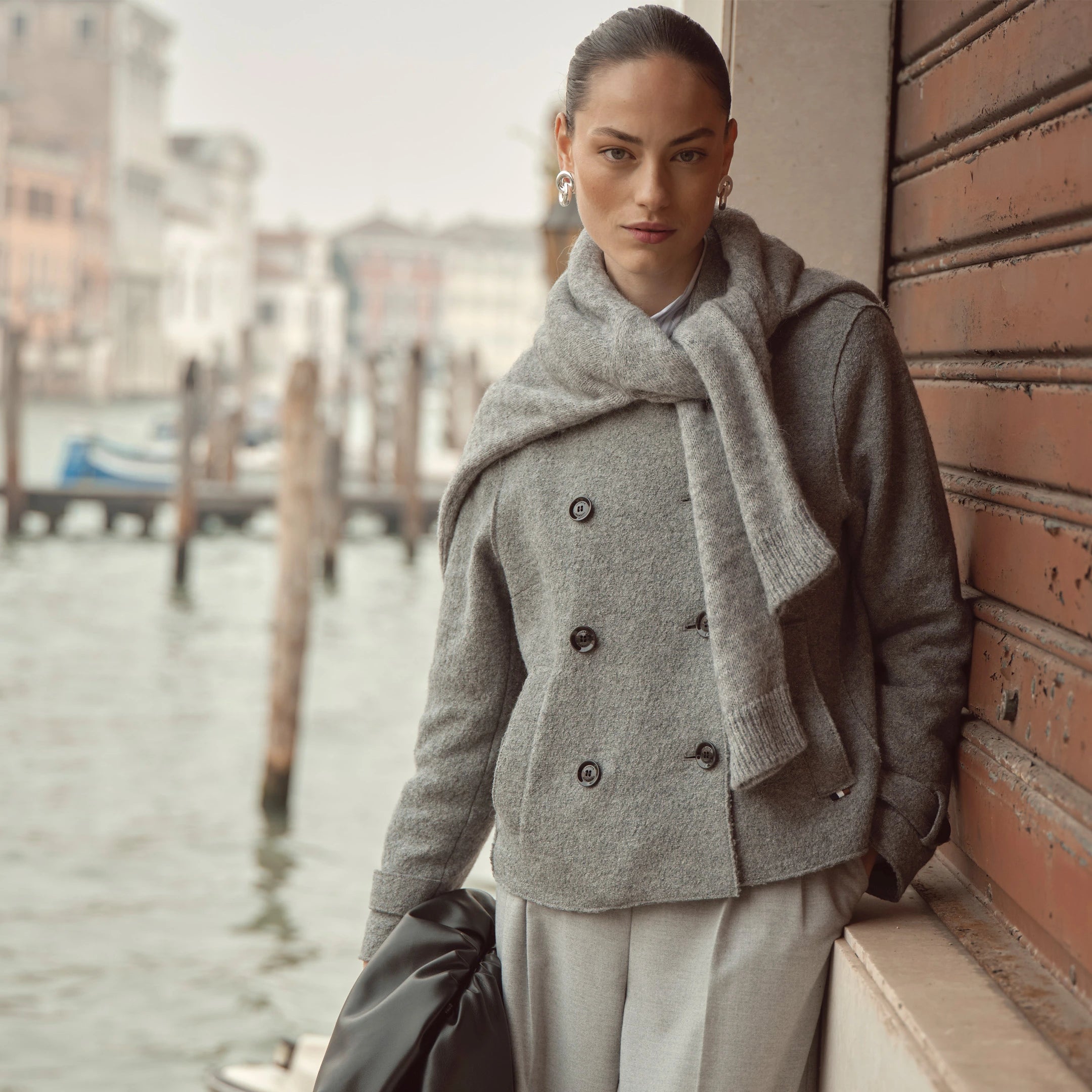 Frau trägt graue bugatti Jacke im Double-Breasted-Stil mit locker über die Schultern gelegtem Strickpullover, kombiniert mit weitem Hosenlook – elegantes Outfit der Damen Herbst/Winter-Kollektion 2025 im Venetian Chic.