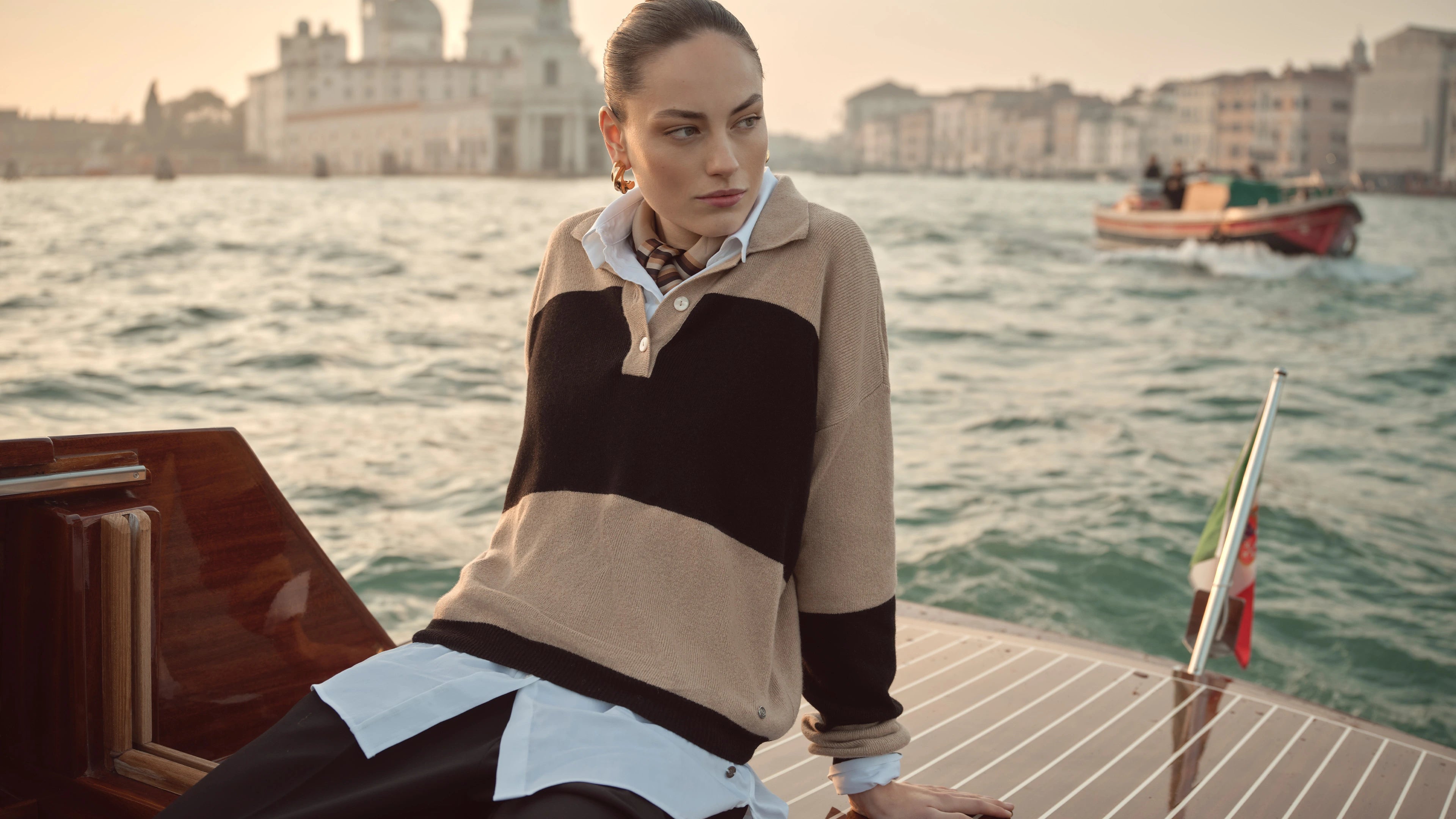 Frau trägt beigefarbenen bugatti Pullover mit schwarzen Blockstreifen über weißer Bluse, aufgenommen auf einem Boot in Venedig – stilvoller Damenlook der Herbst/Winter-Kollektion 2025 im Venetian Chic.