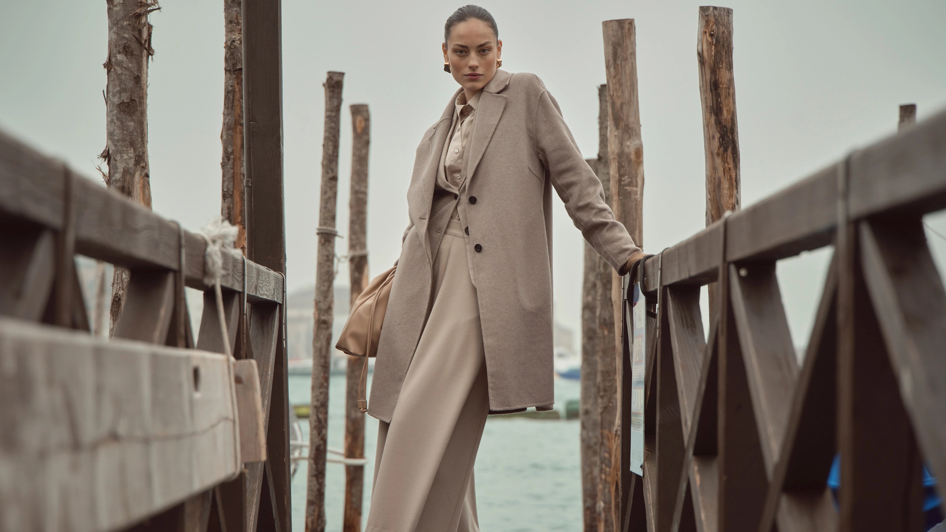 Frau trägt beigen bugatti-Mantel mit eleganter Bluse und weiter Hose in Ton-in-Ton-Optik, kombiniert mit Tasche – stilvoller Look am Wasser in Venedig.