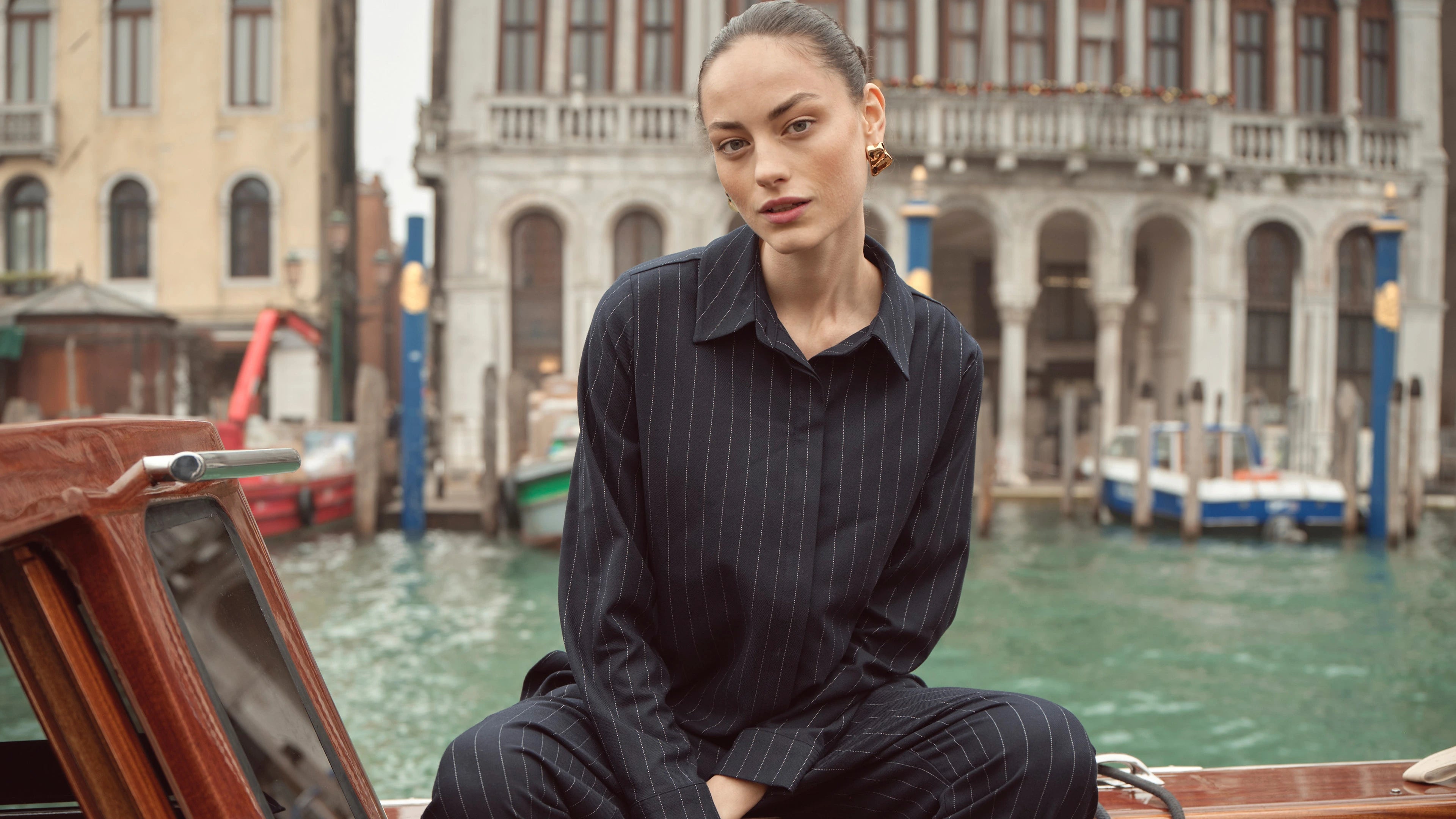Frau sitzt in dunkelblauem Nadelstreifen-Ensemble von bugatti auf einem Boot vor venezianischer Kulisse – eleganter Damenlook der Herbst/Winter-Kollektion 2025 mit moderner Business-Note.