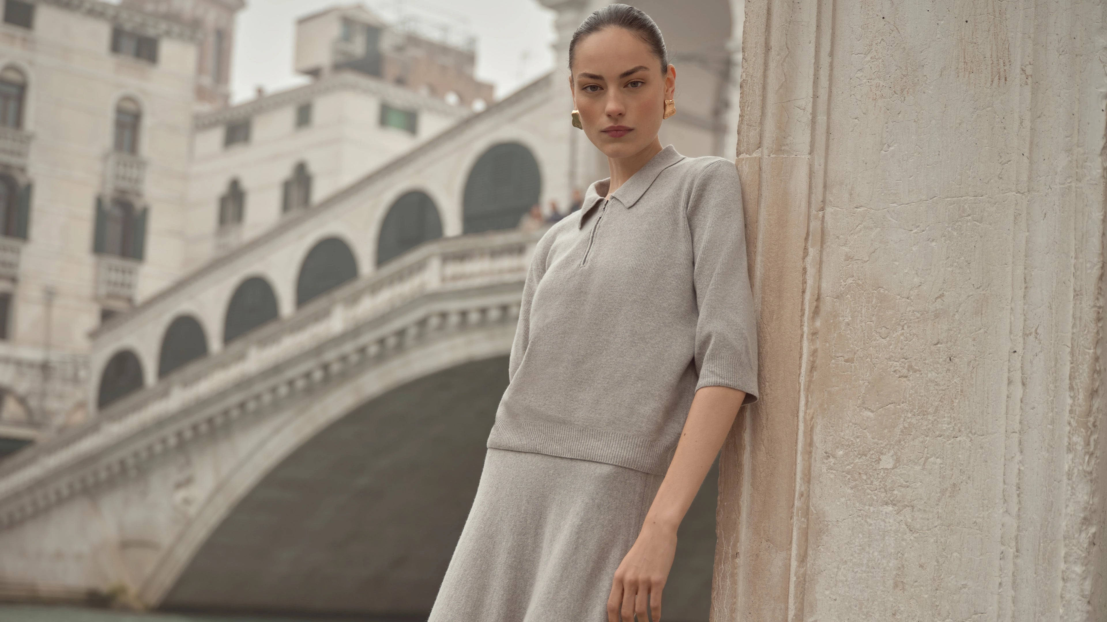 Frau in grauem bugatti-Strick-Set mit halblangem Ärmelpullover und Rock, elegant inszeniert vor der Rialto-Brücke in Venedig.