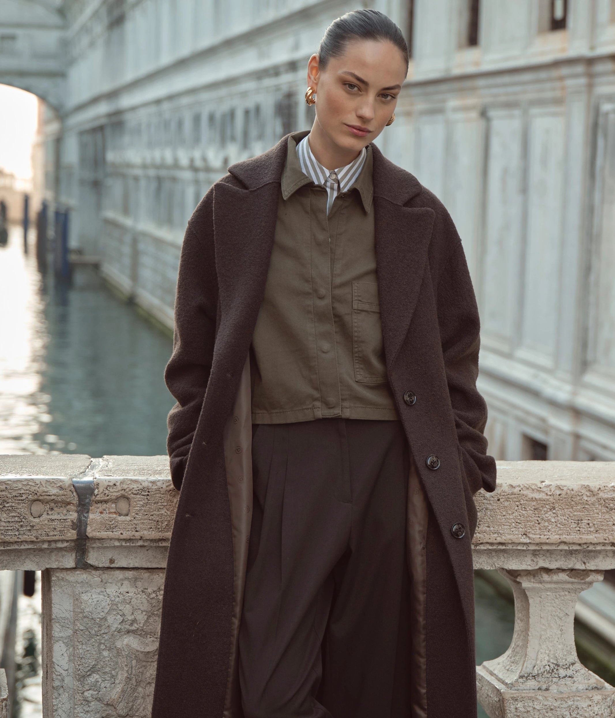 Frau in elegantem bugatti-Look mit langem braunen Mantel und olivfarbener Bluse, fotografiert vor historischer Architektur in Venedig.