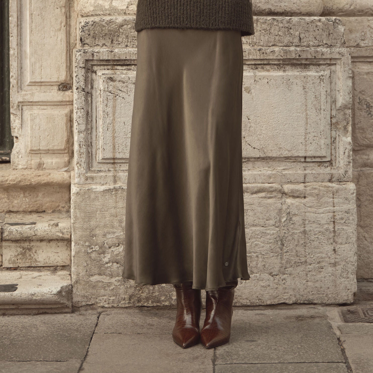 Frau trägt olivfarbenen bugatti Midirock aus fließendem Satin, kombiniert mit braunen Stiefeletten – eleganter Look der Damen Herbst/Winter-Kollektion 2025 im Venetian Chic.