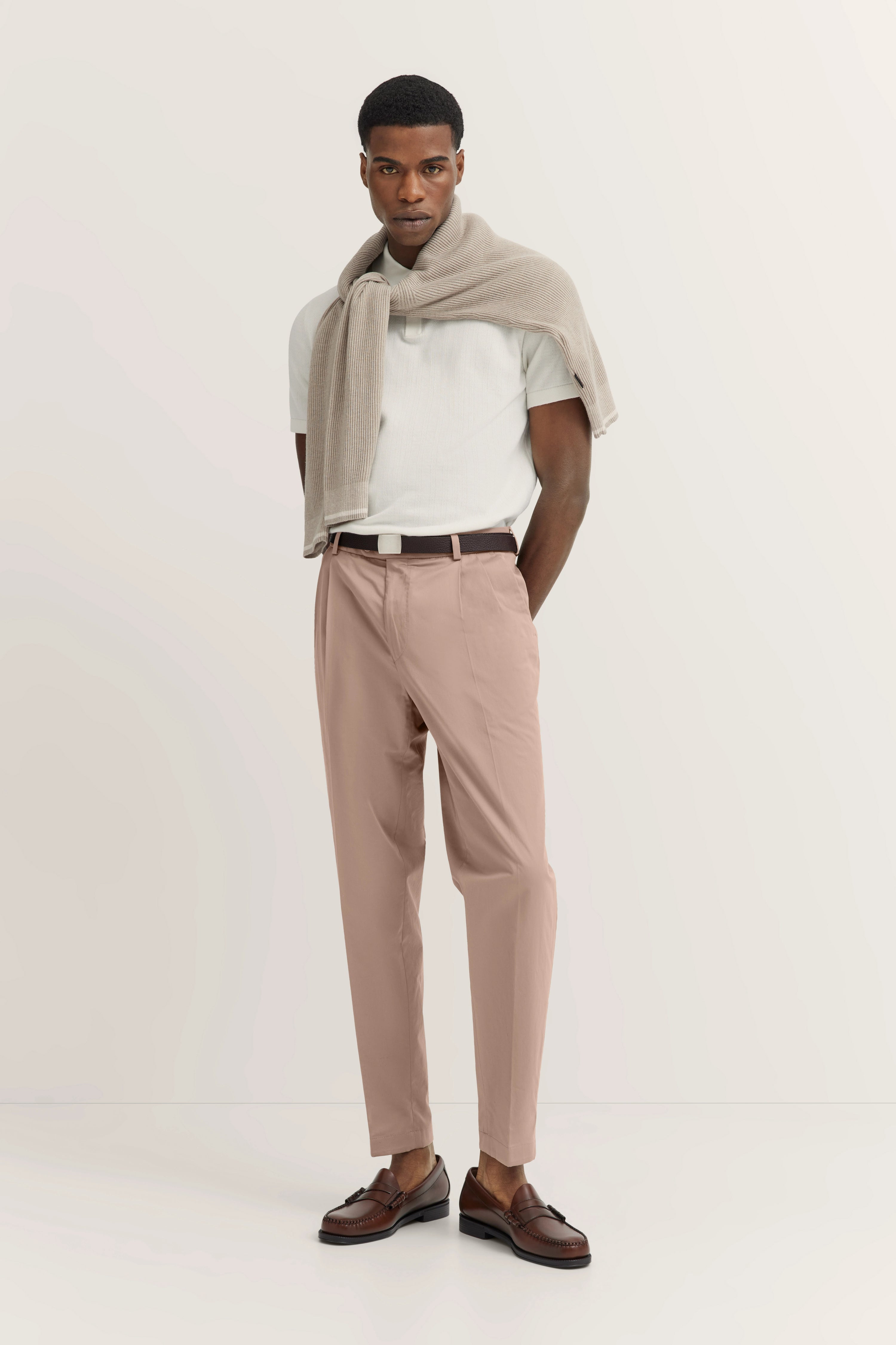 Model in beige trui over wit poloshirt, roze chino, bruine loafers. Ontspannen, zelfverzekerd.