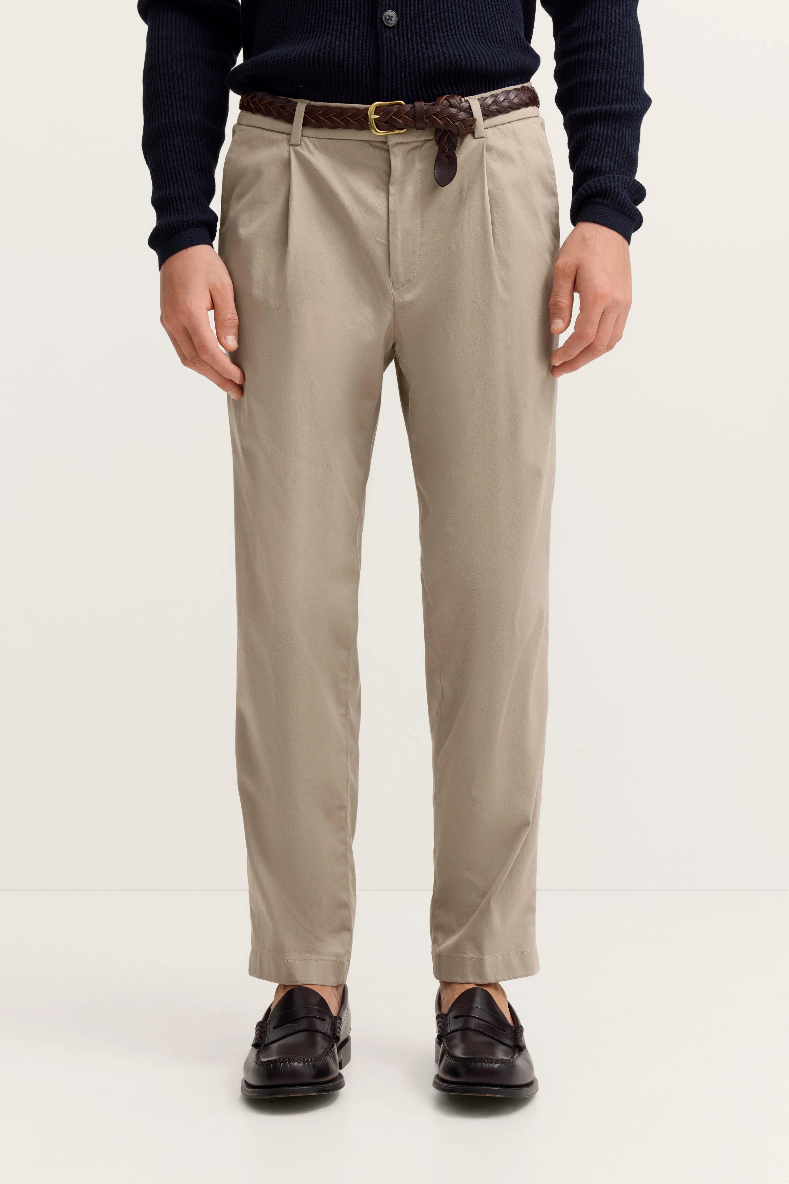 Beige Chinos mit braunem Ledergürtel, Modelbild, entspannte Pose, schwarze Loafer.