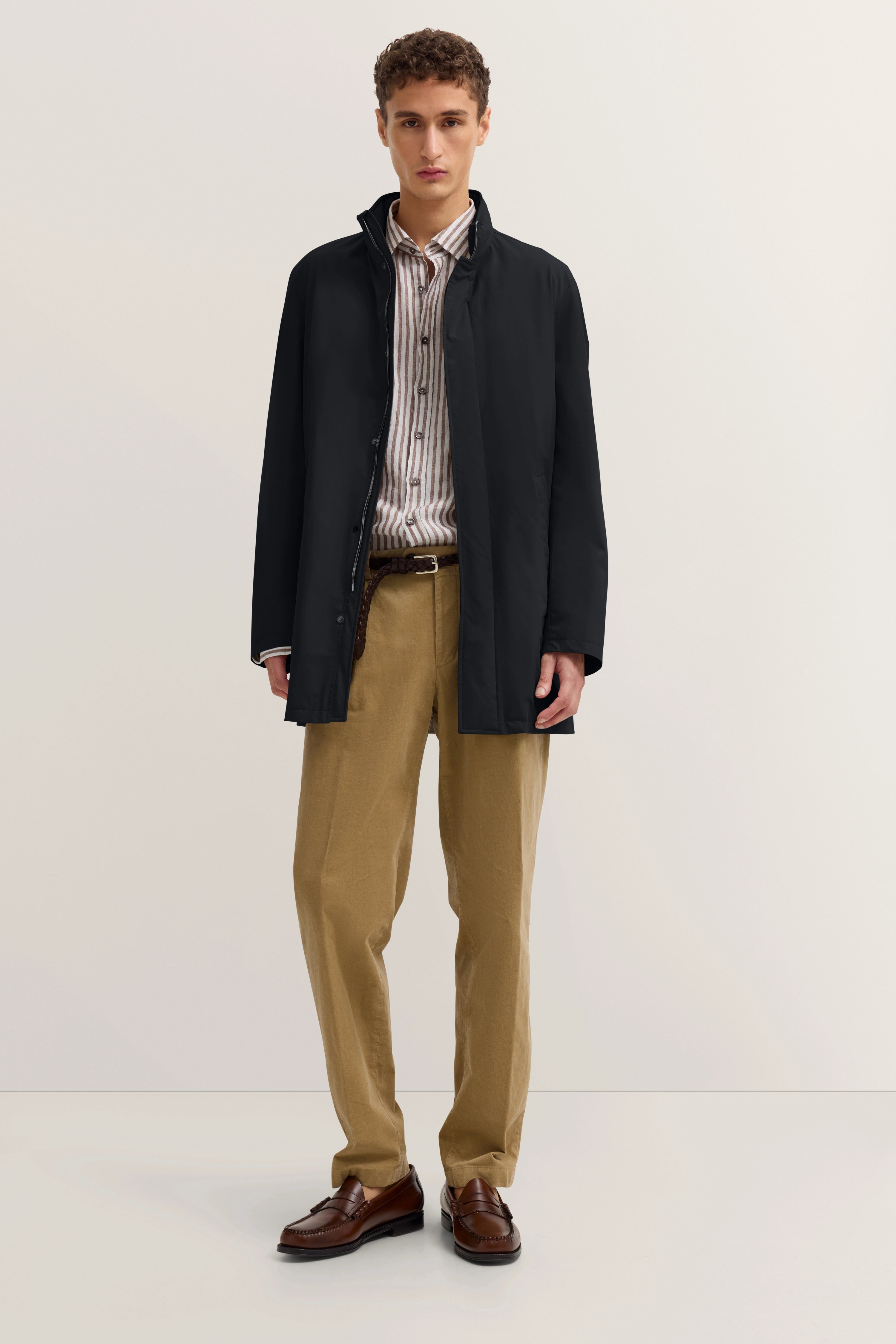 Model in schwarzem Mantel, gestreiftem Hemd, beigen Hosen und braunen Schuhen, frontal stehend.