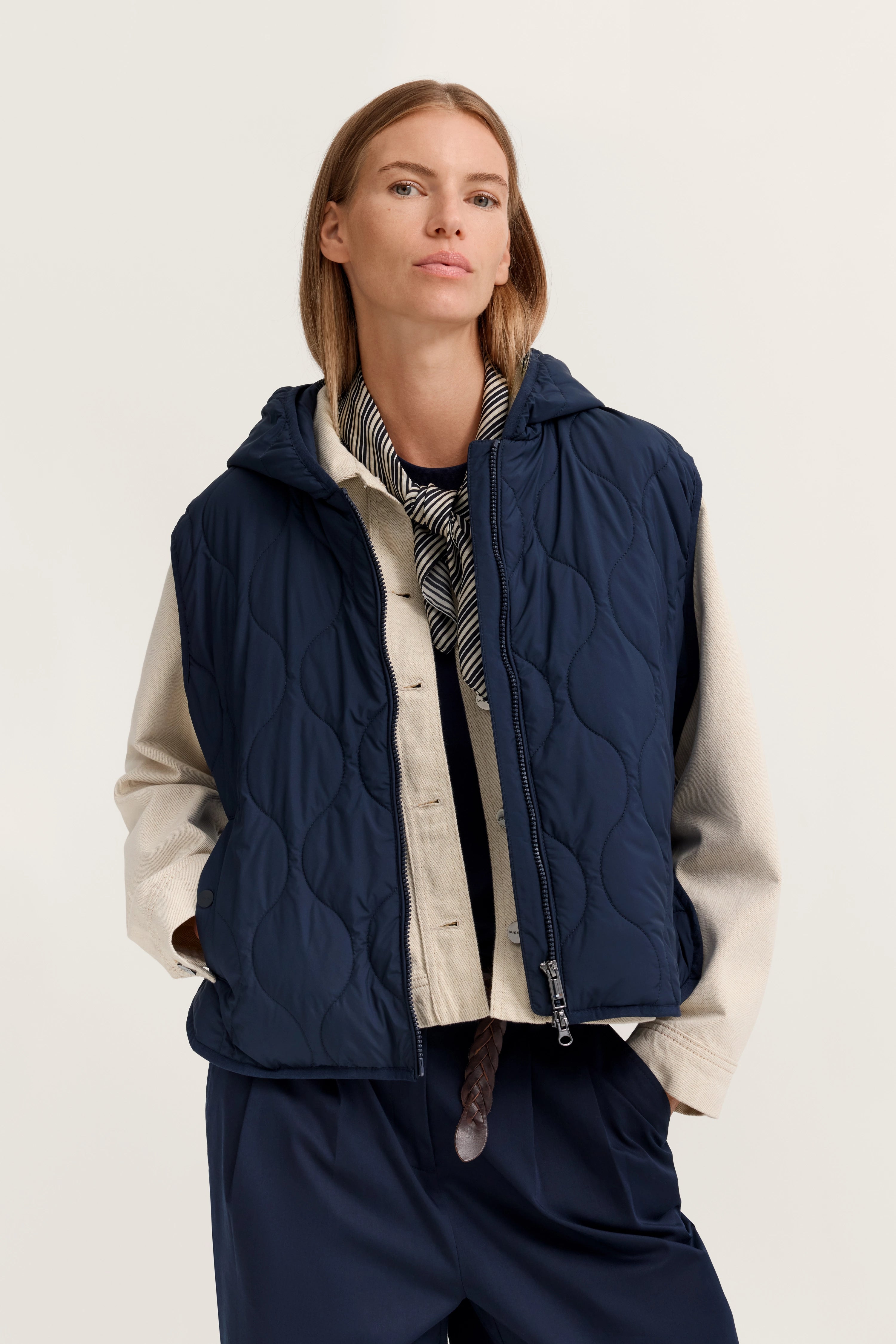 Modelbild: Frau trägt blaue Steppweste, beige Jacke, gestreiftes Tuch. Lässig, Hände in Taschen.