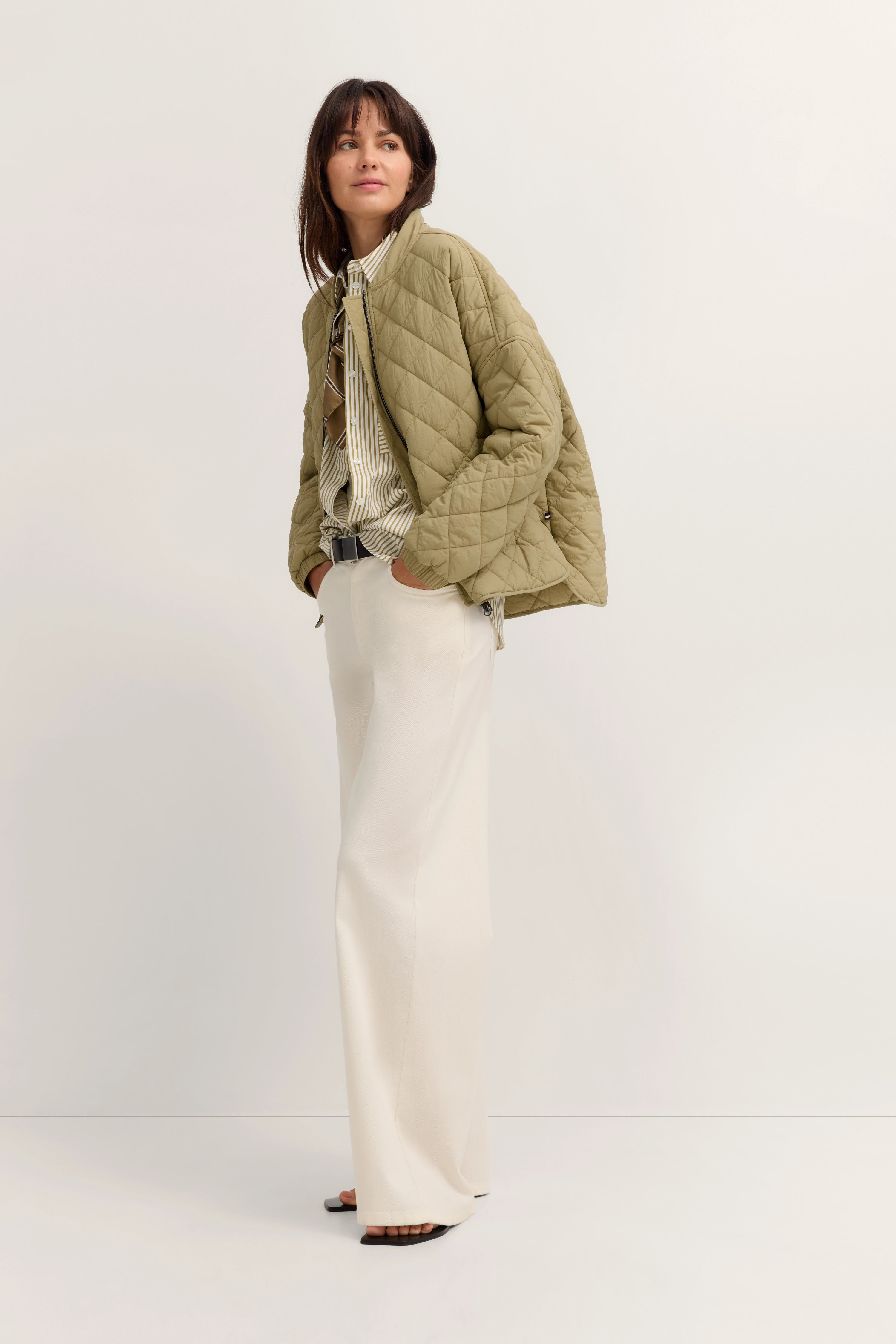 Model im legeren Outfit mit olivgrüner Steppjacke, gestreiftem Hemd und weißen Hosen.