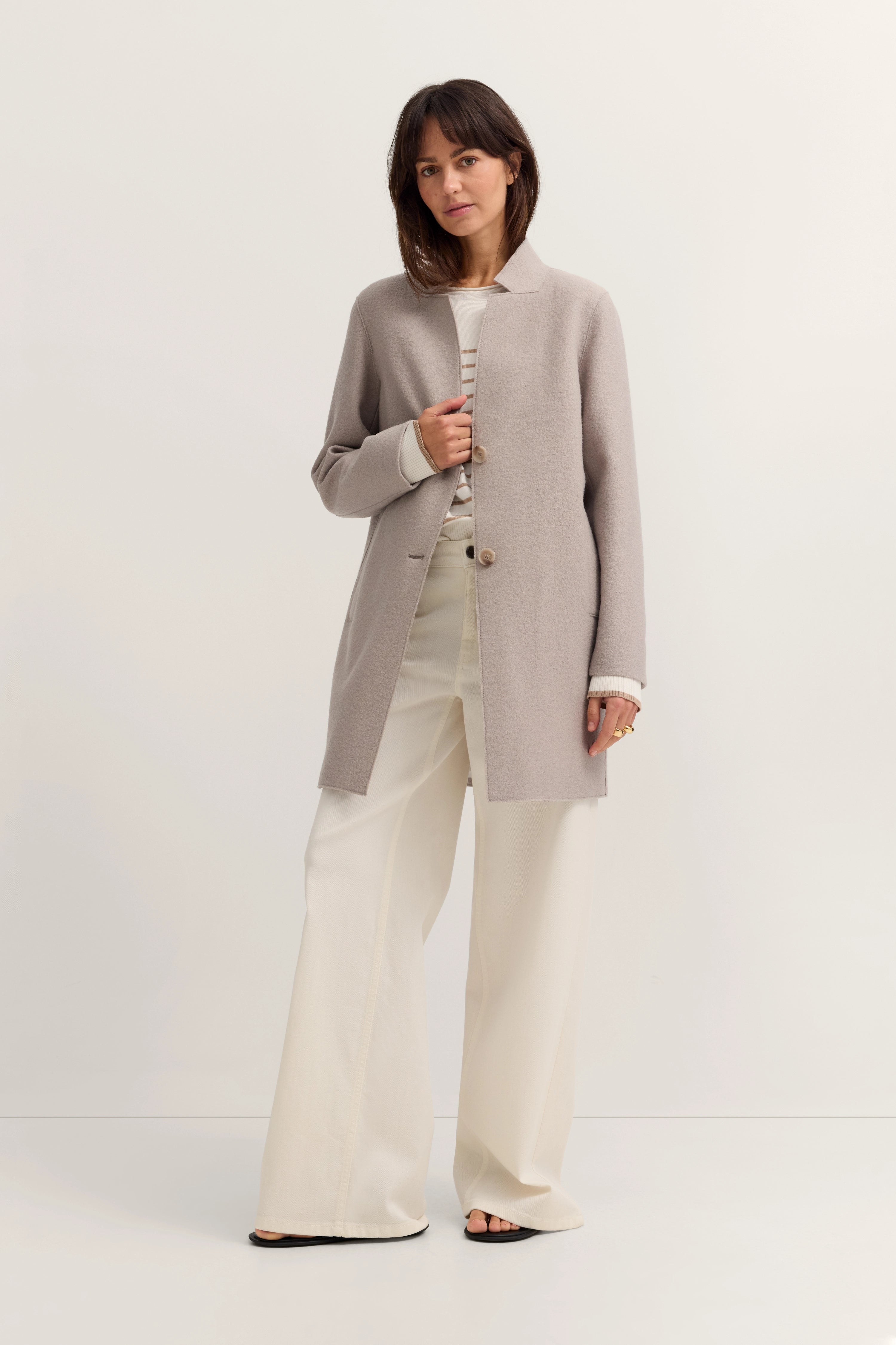 Model mit beige bugatti Mantel, weißem Top und weiter Hose, entspannte Pose.