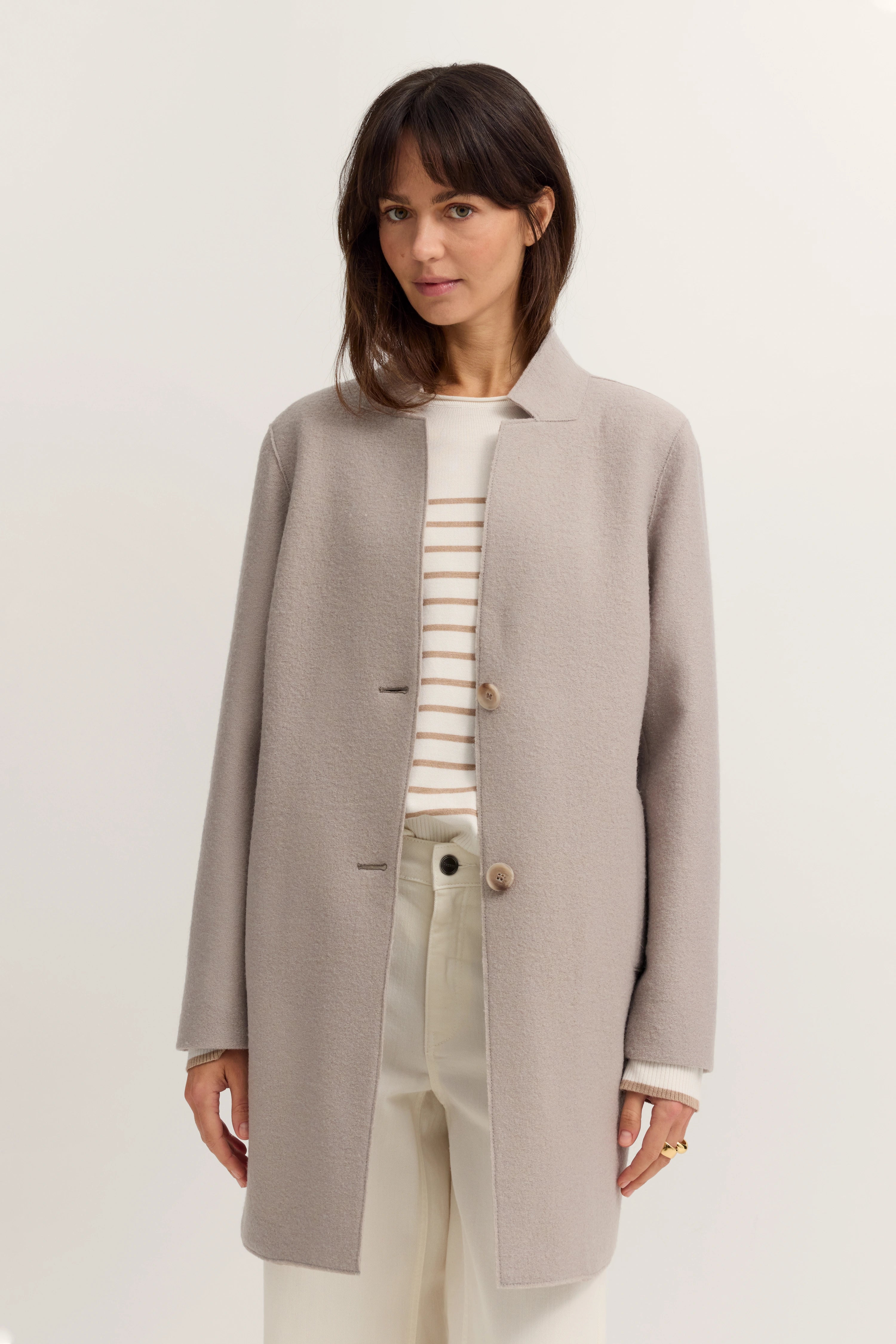 Grauer Mantel über gestreiftem Pullover, creme Hose, Modelbild, entspanntes Posing.