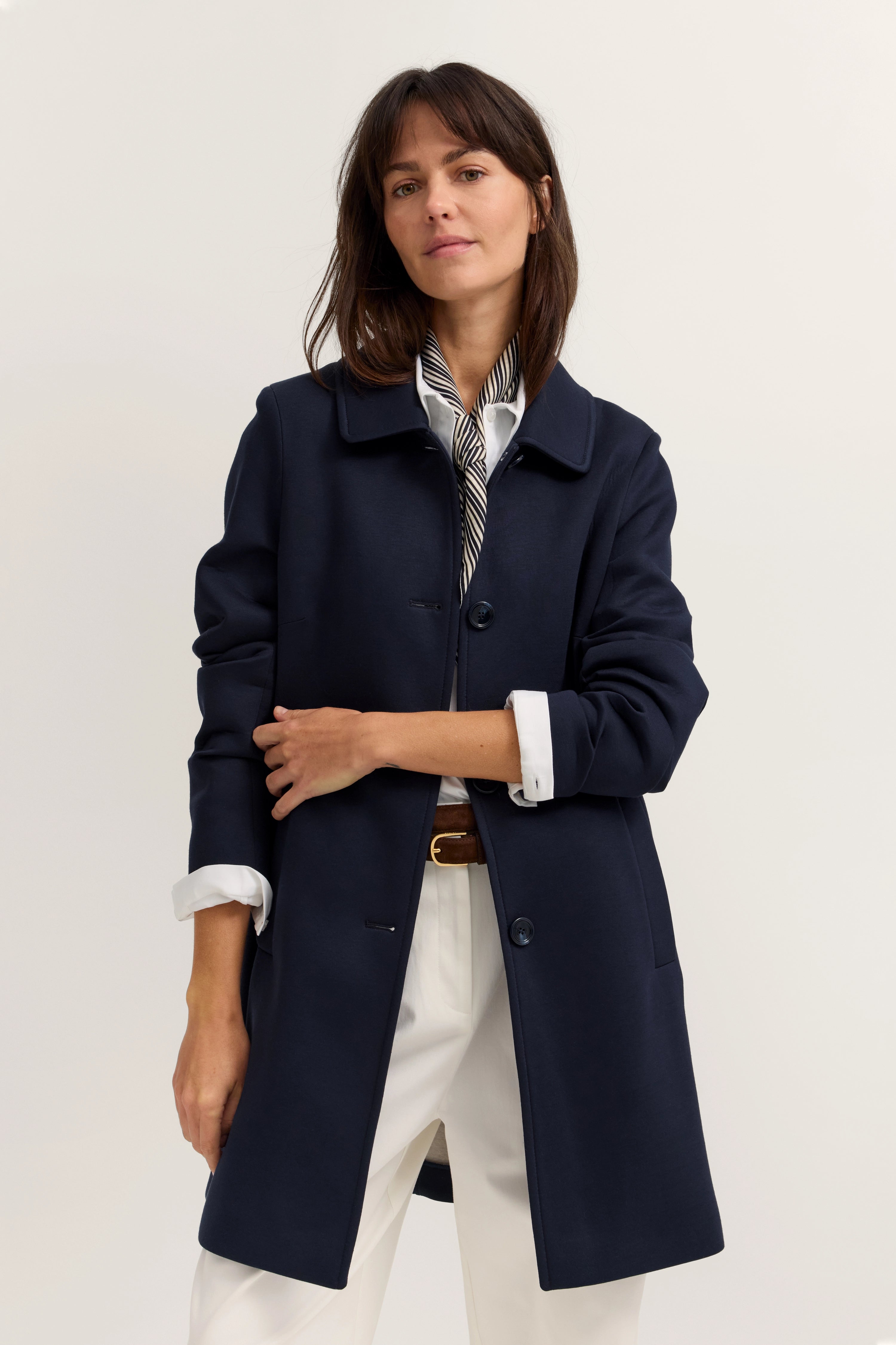 Damenoutfit mit marineblauem Mantel, weißer Hose, gestreiftem Schal, Model posiert mit verschränkten Armen.