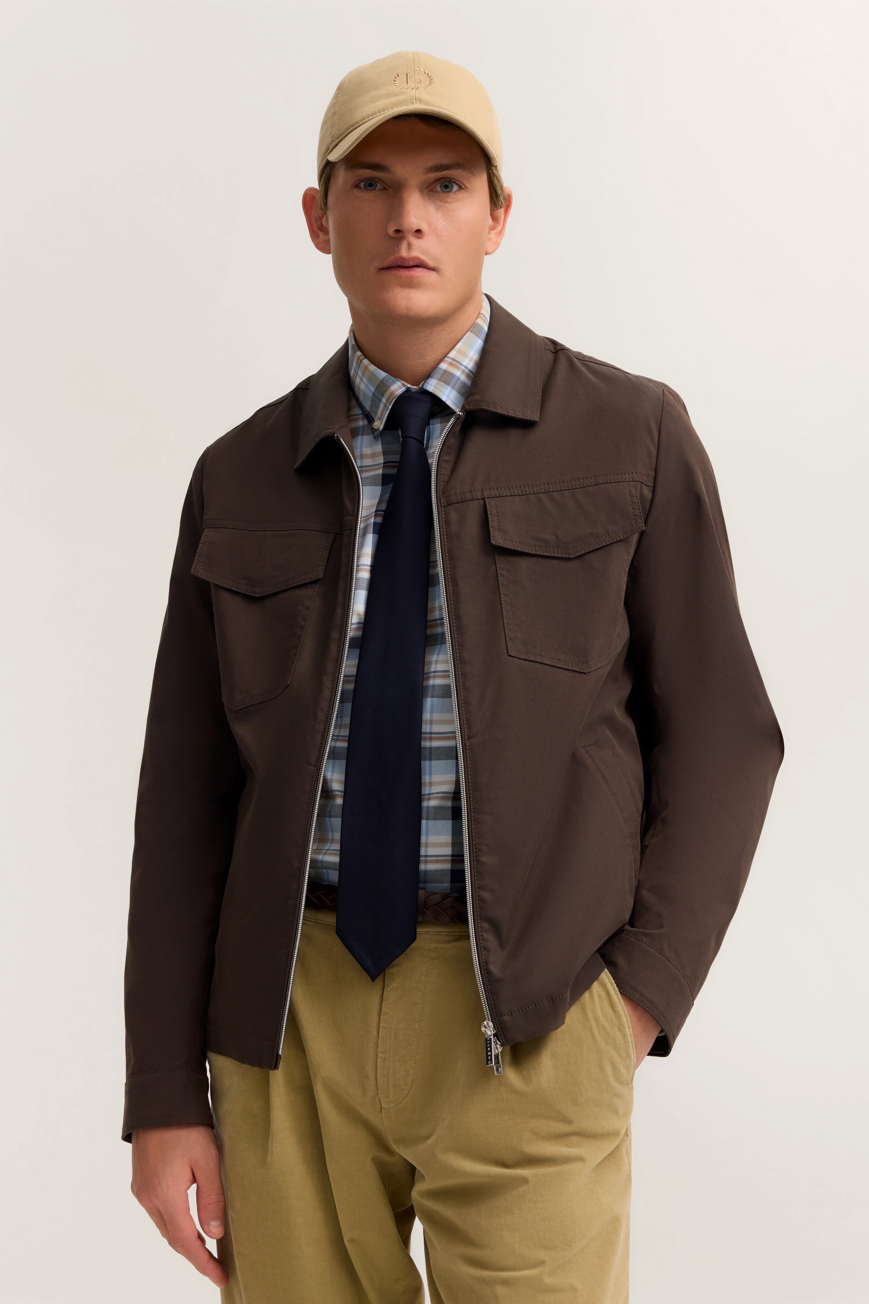 Model in brauner Jacke, karierter Hemd, dunkelblauer Krawatte, beige Hose. Lässig mit beiger Kappe.