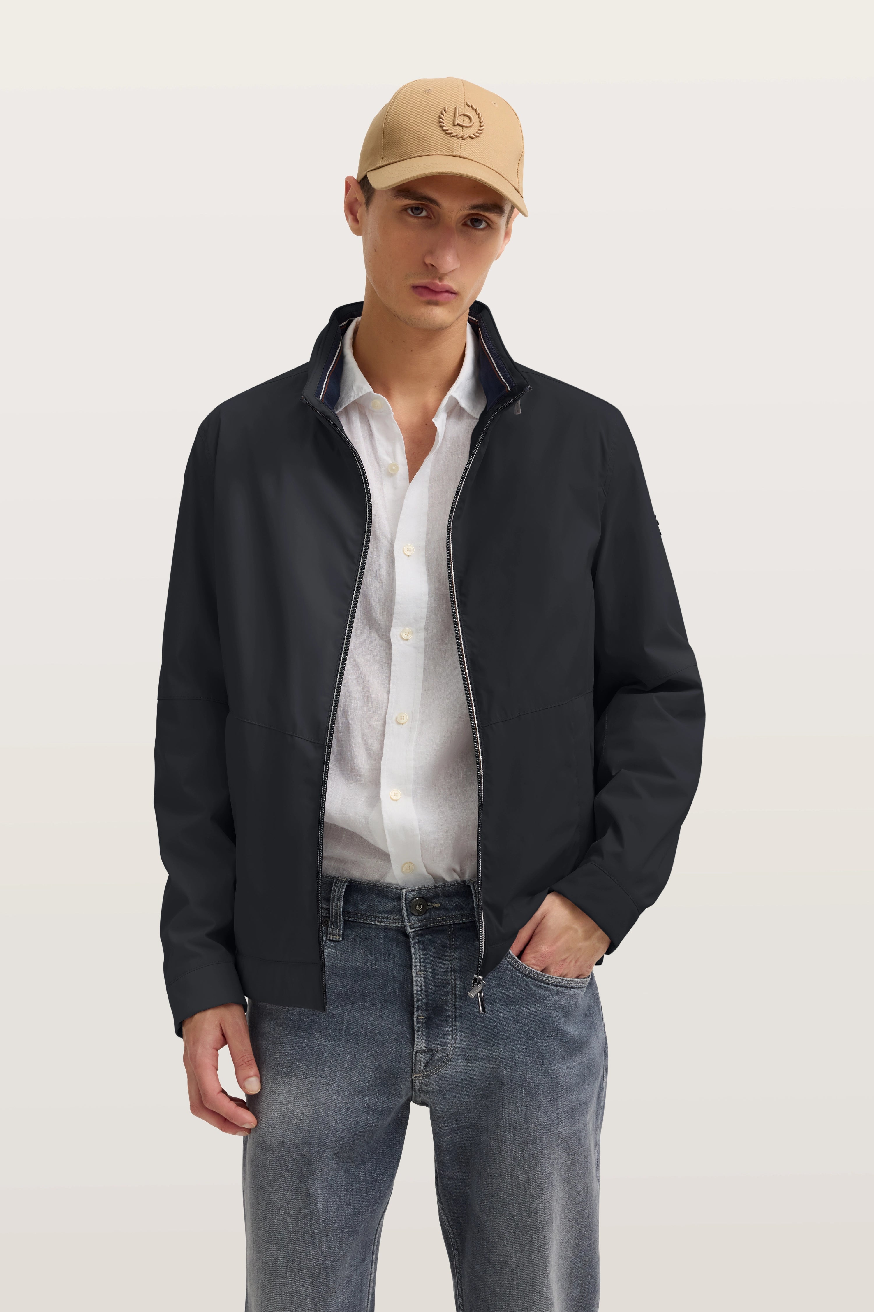 Model trägt schwarze bugatti Jacke, weißes Hemd, Jeans und beige Kappe, lässige Pose.