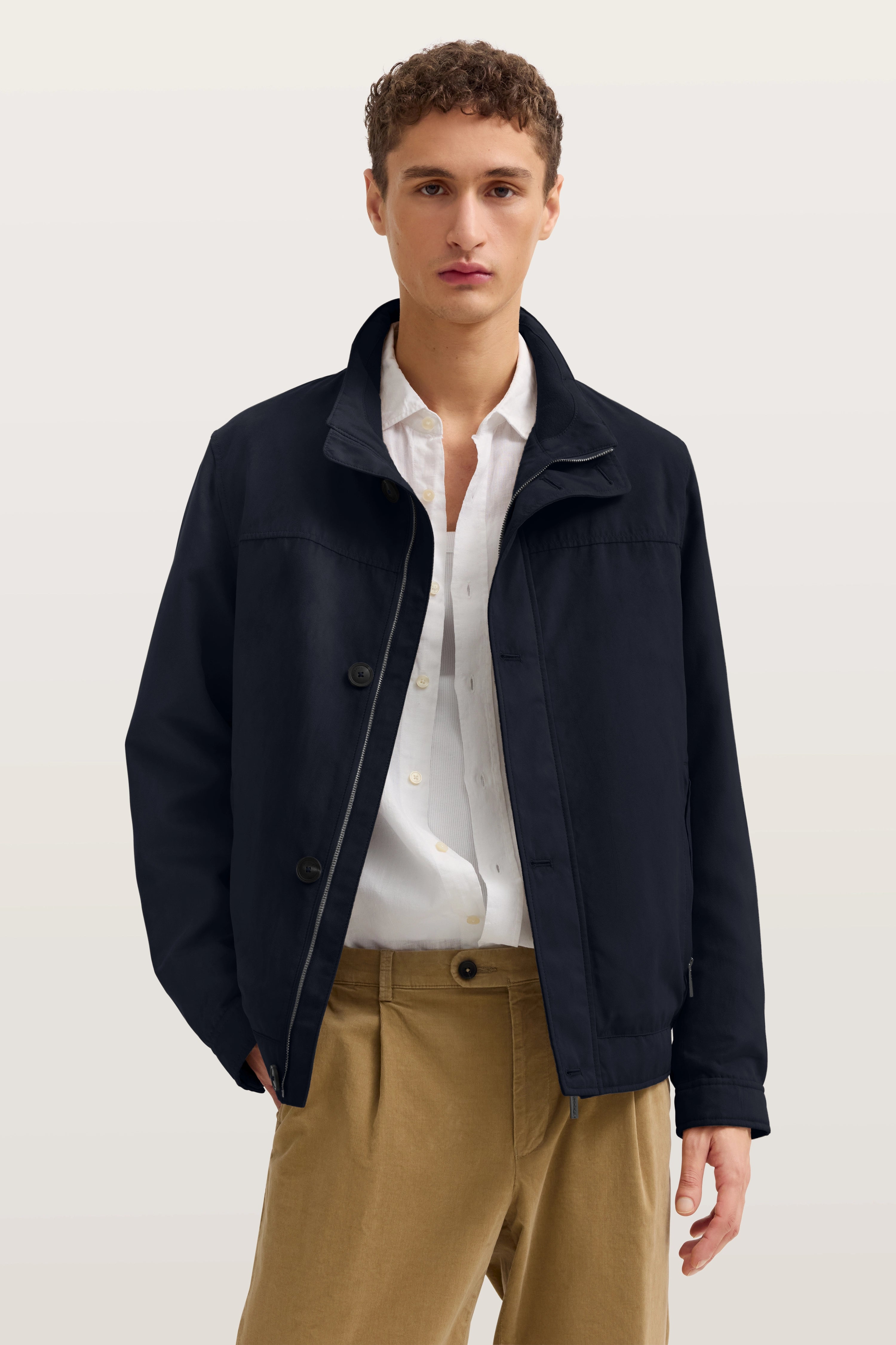 Model in beiger Hose, weißem Hemd und dunkelblauer bugatti Jacke, frontal stehend.