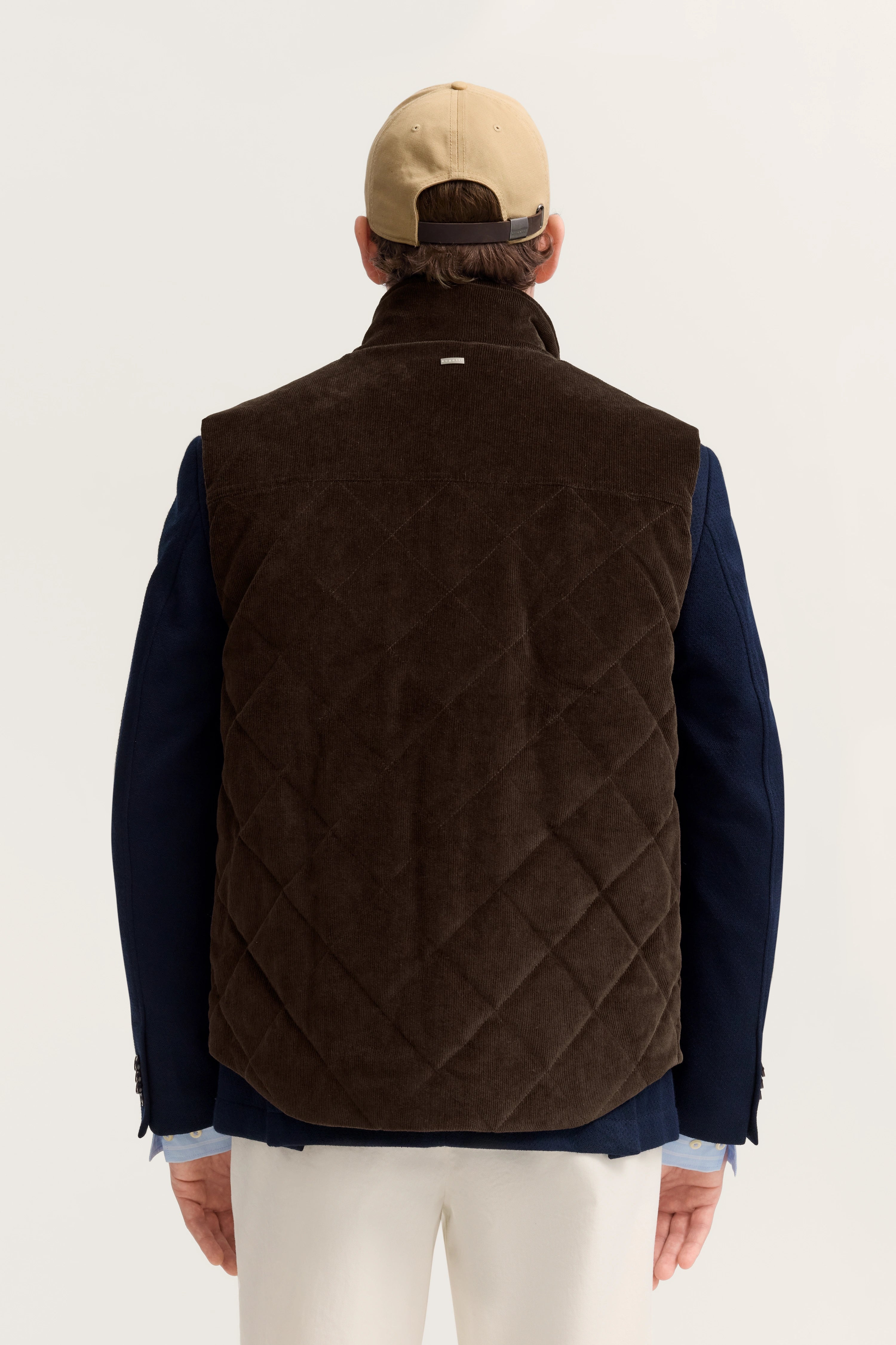 Rückansicht Model in brauner Steppweste, blauem Sakko, weißer Hose, beigefarbene Cap.