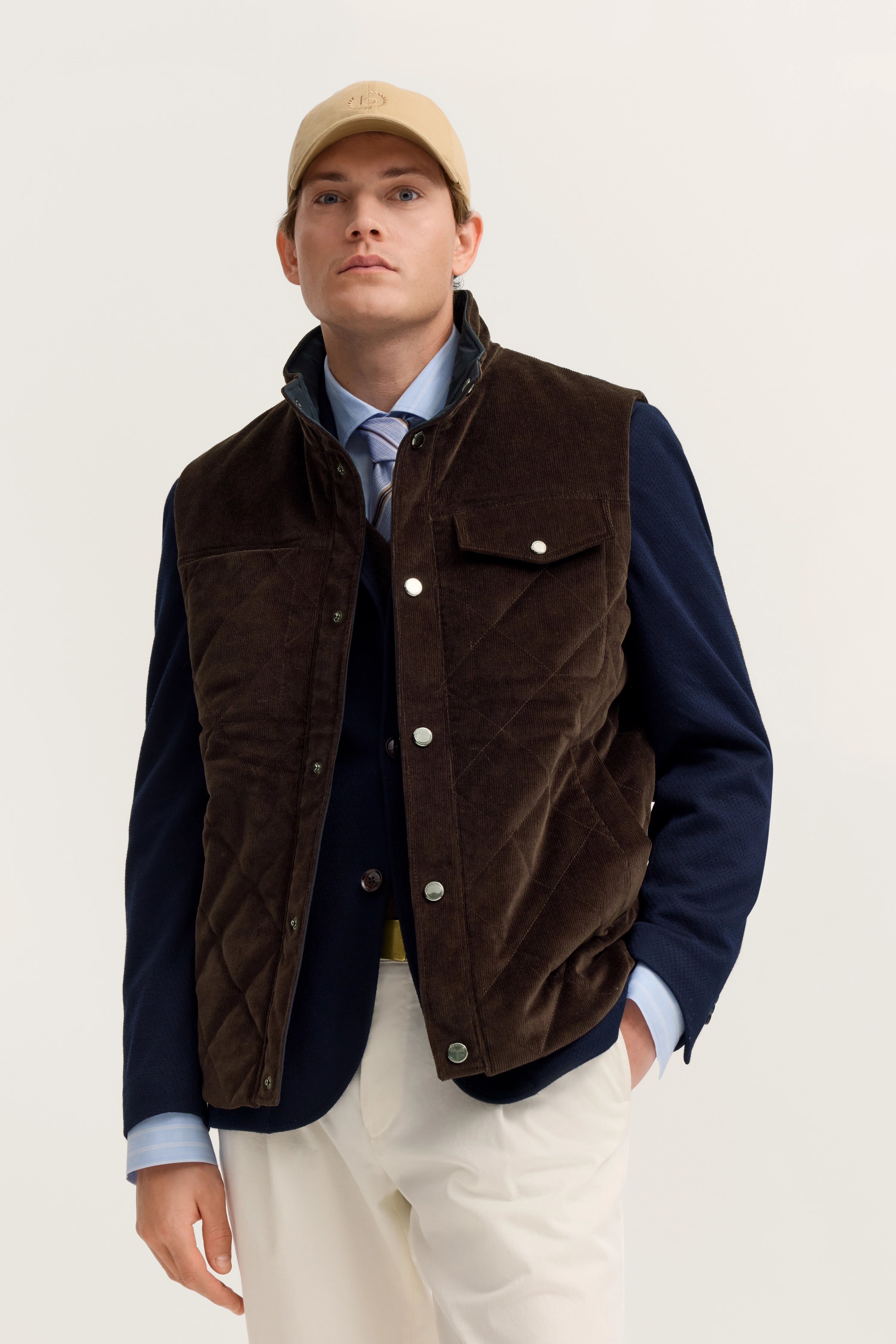 Model in brauner Weste, blauem Blazer und beiger Hose. Legere Pose, cap ergänzt den Look.