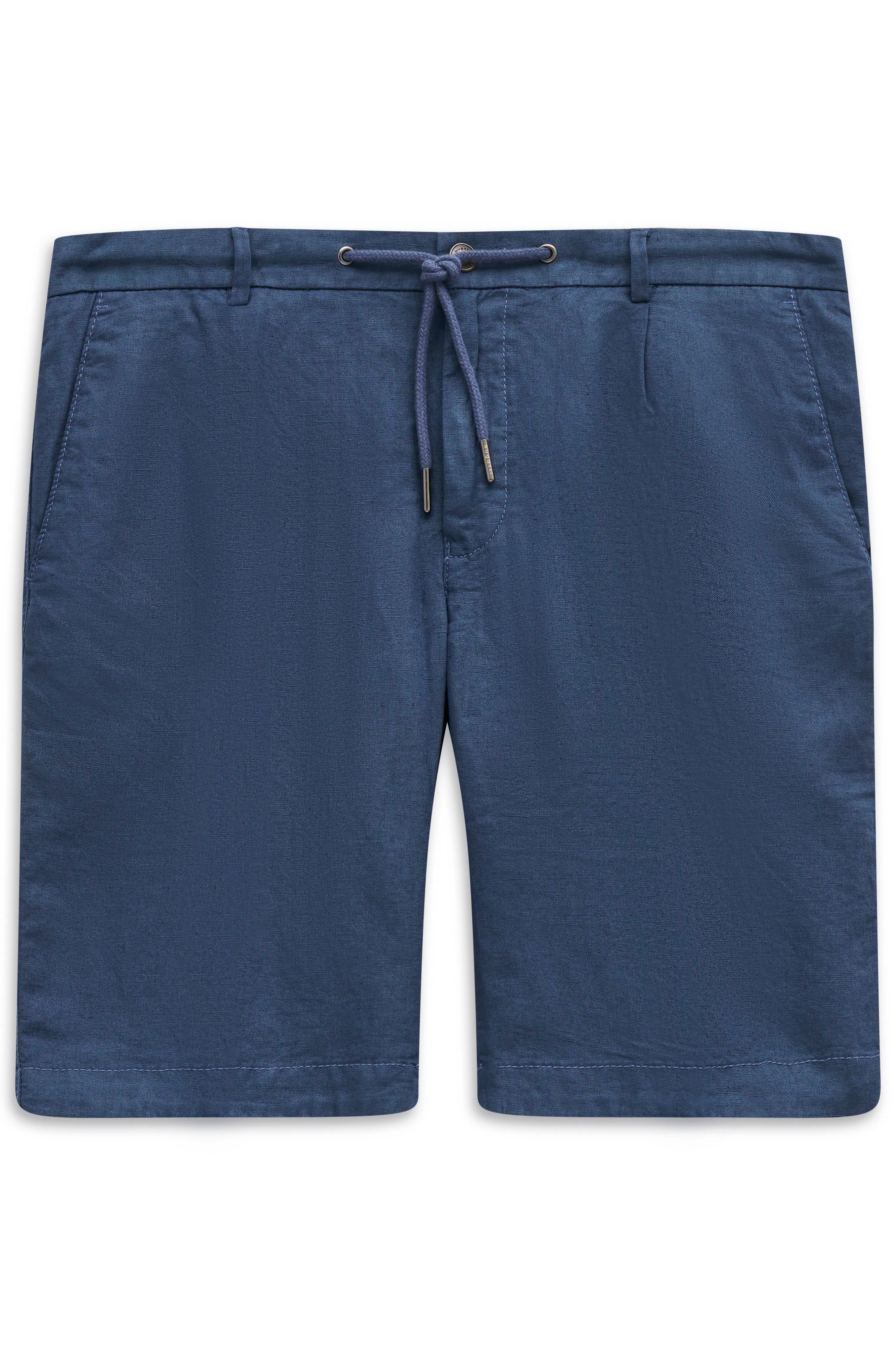 Model in weißem Hemd, blauen Shorts, beigem Pullover über Schultern, gemusterte Tasche, schwarze Sandalen.