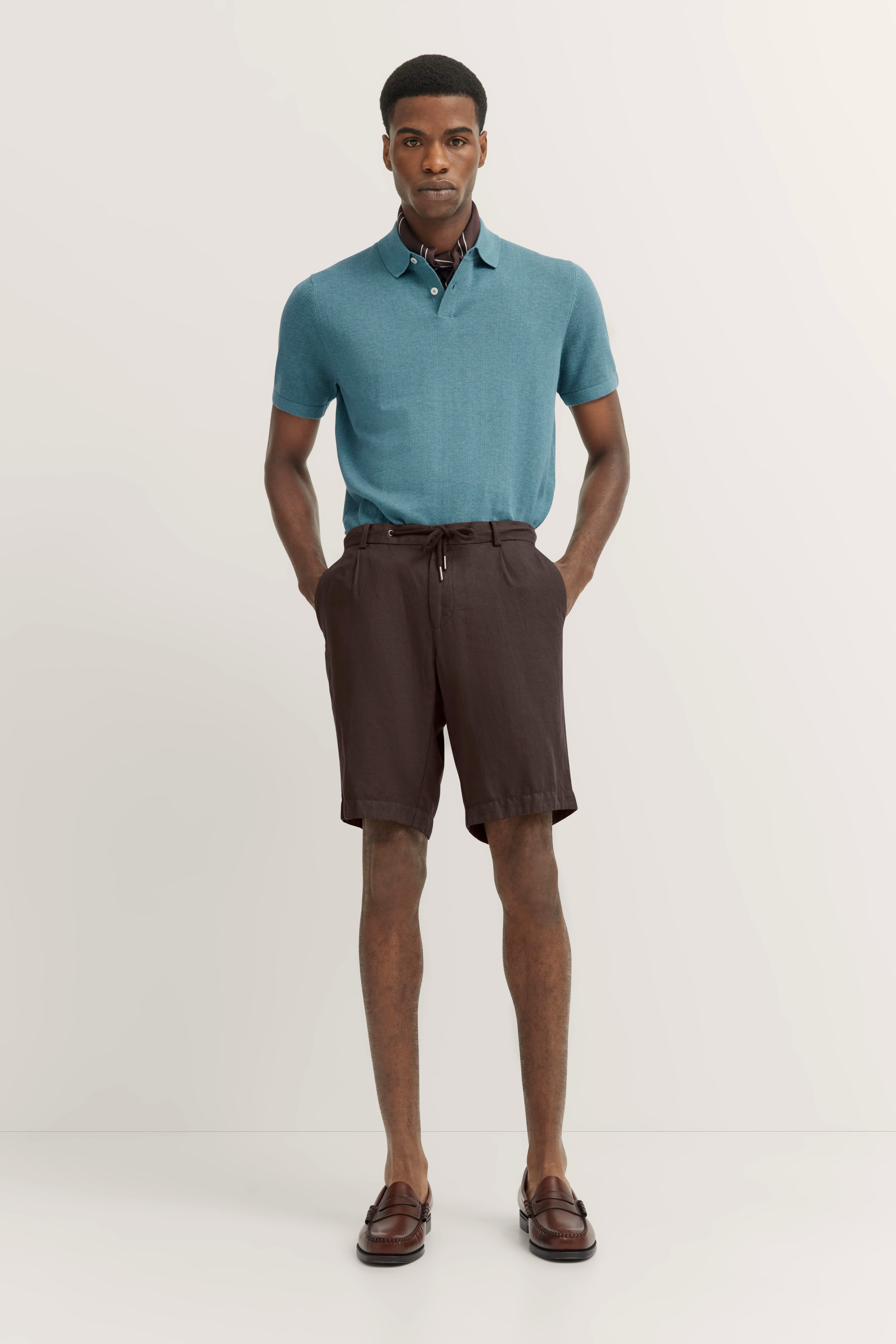 Model in turquoise polo, brown shorts, and loafers, striking a standing pose.