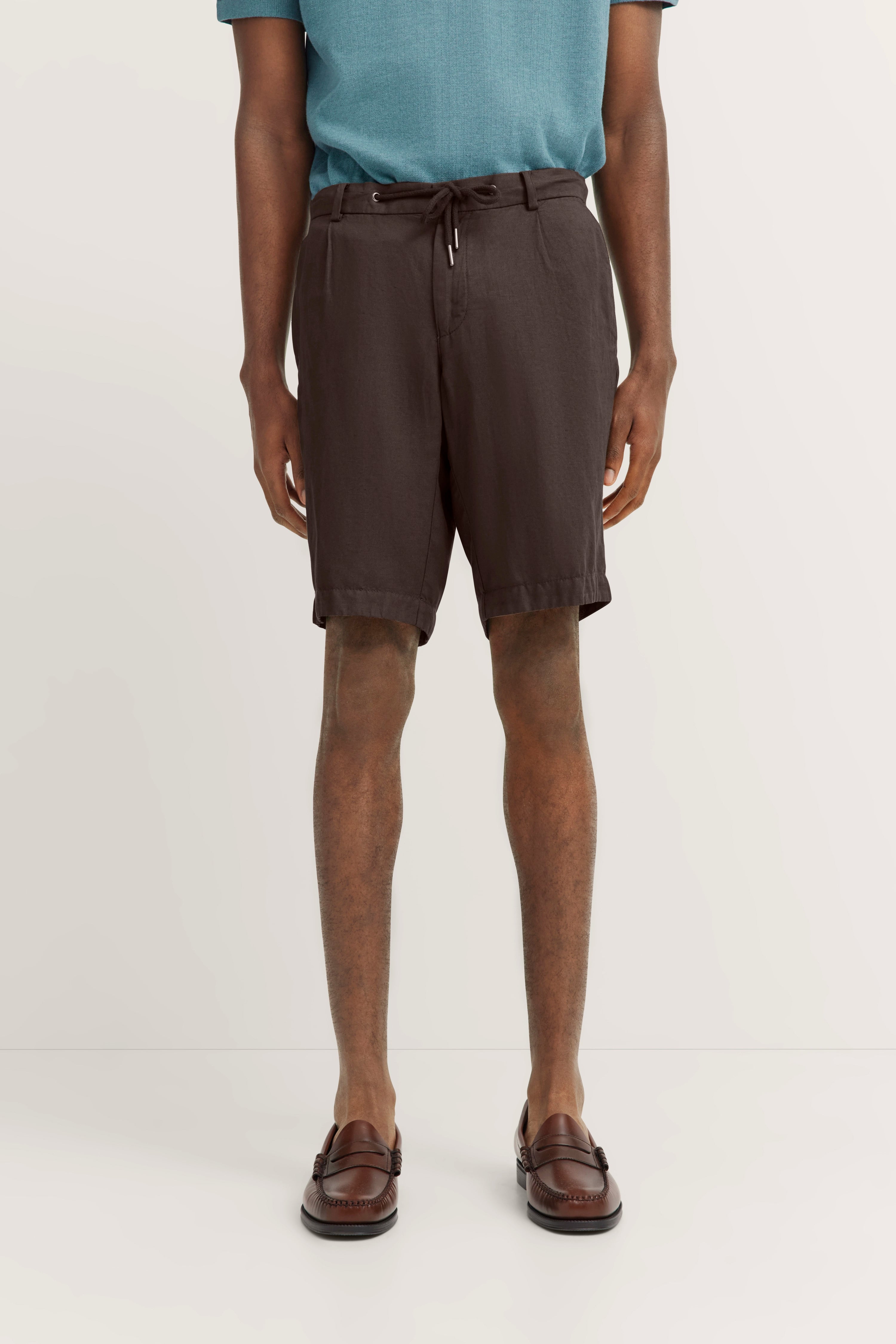 Braunes Shorts-Outfit mit blauem Oberteil und braunen Loafers, legeres Modelbild, entspannte Pose.