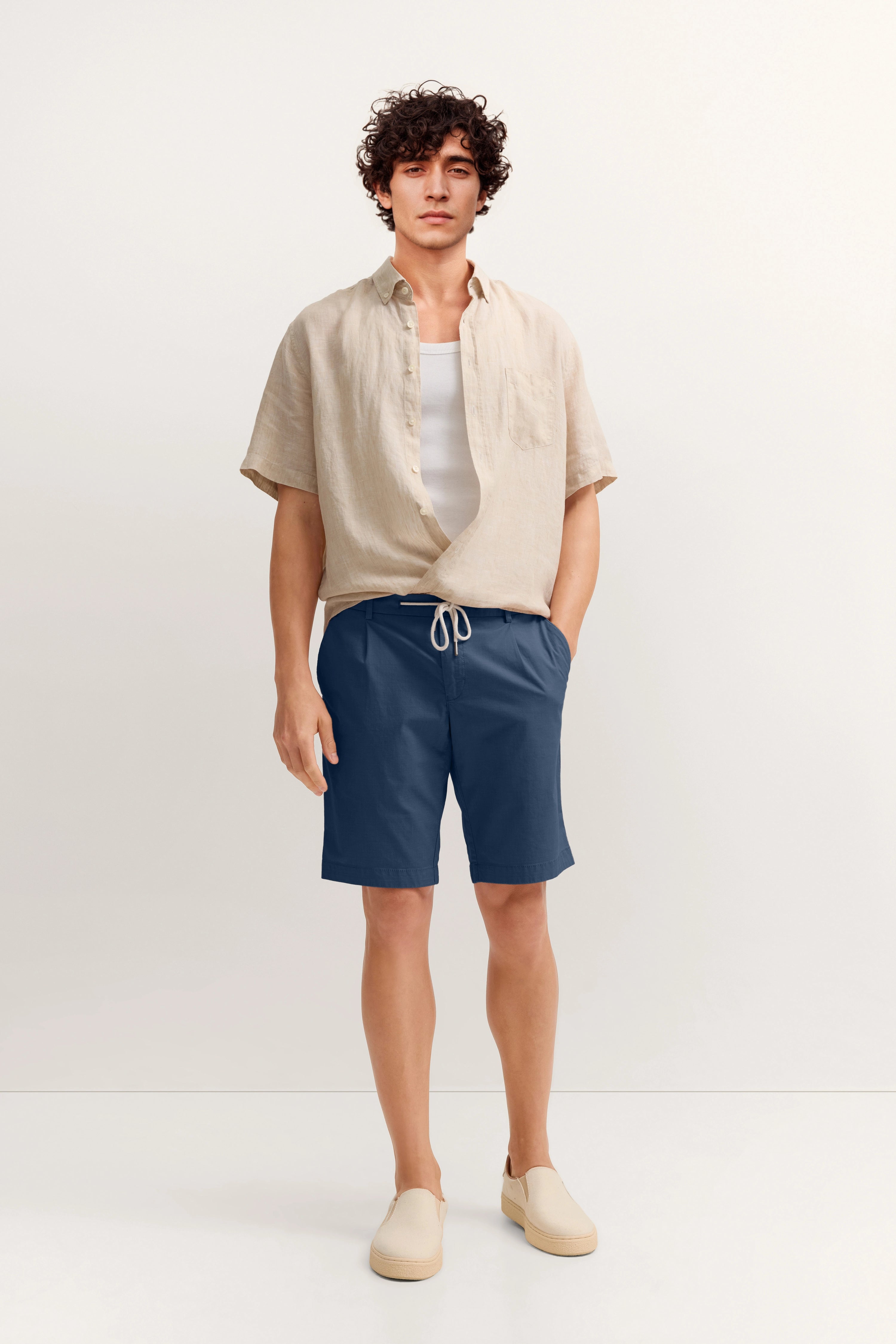 Model im legeren Outfit mit beiger Leinenhemd, blauer Shorts und beigen Schuhen von bugatti.