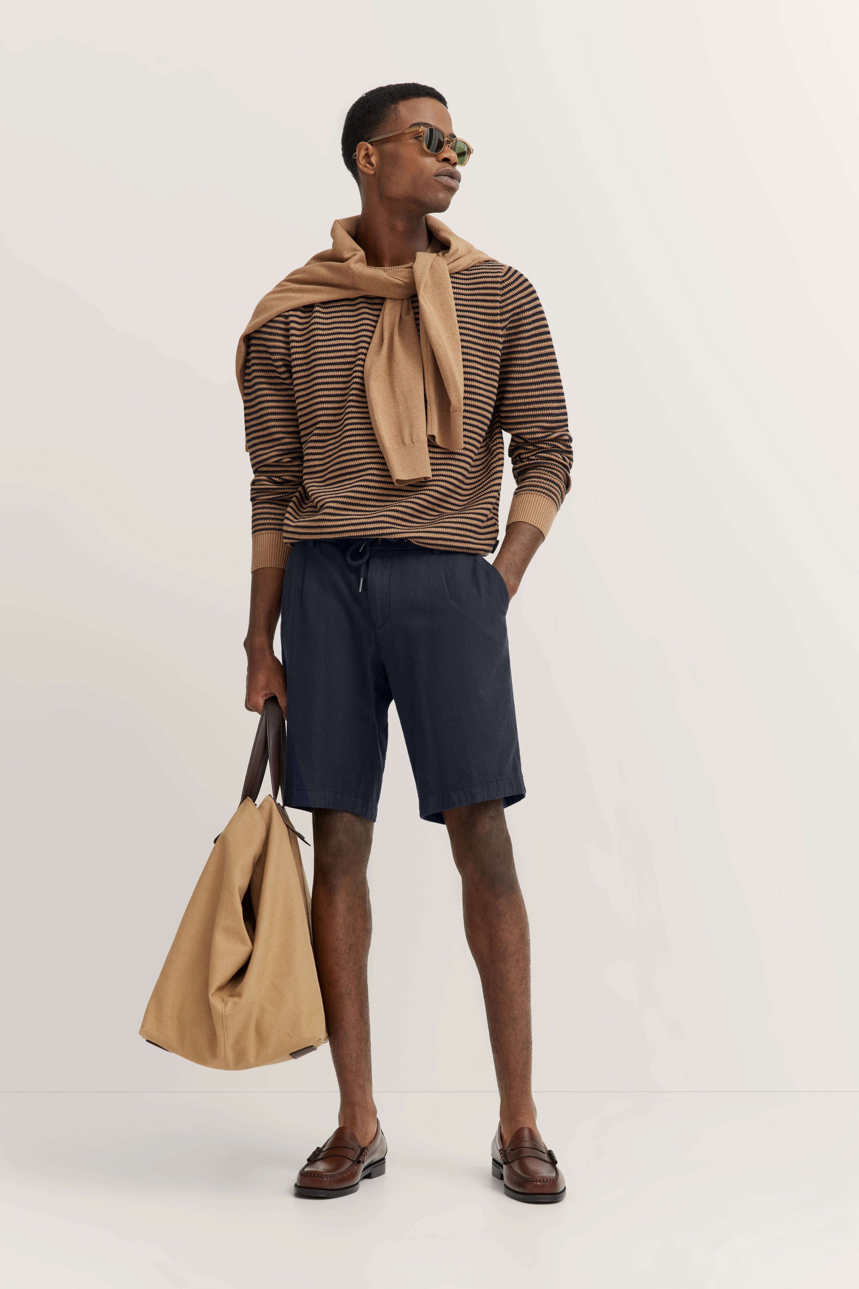 Model in gestreiftem Pullover, blauen Shorts, beigem Pullover um Schultern, lässige Pose, braune Schuhe.