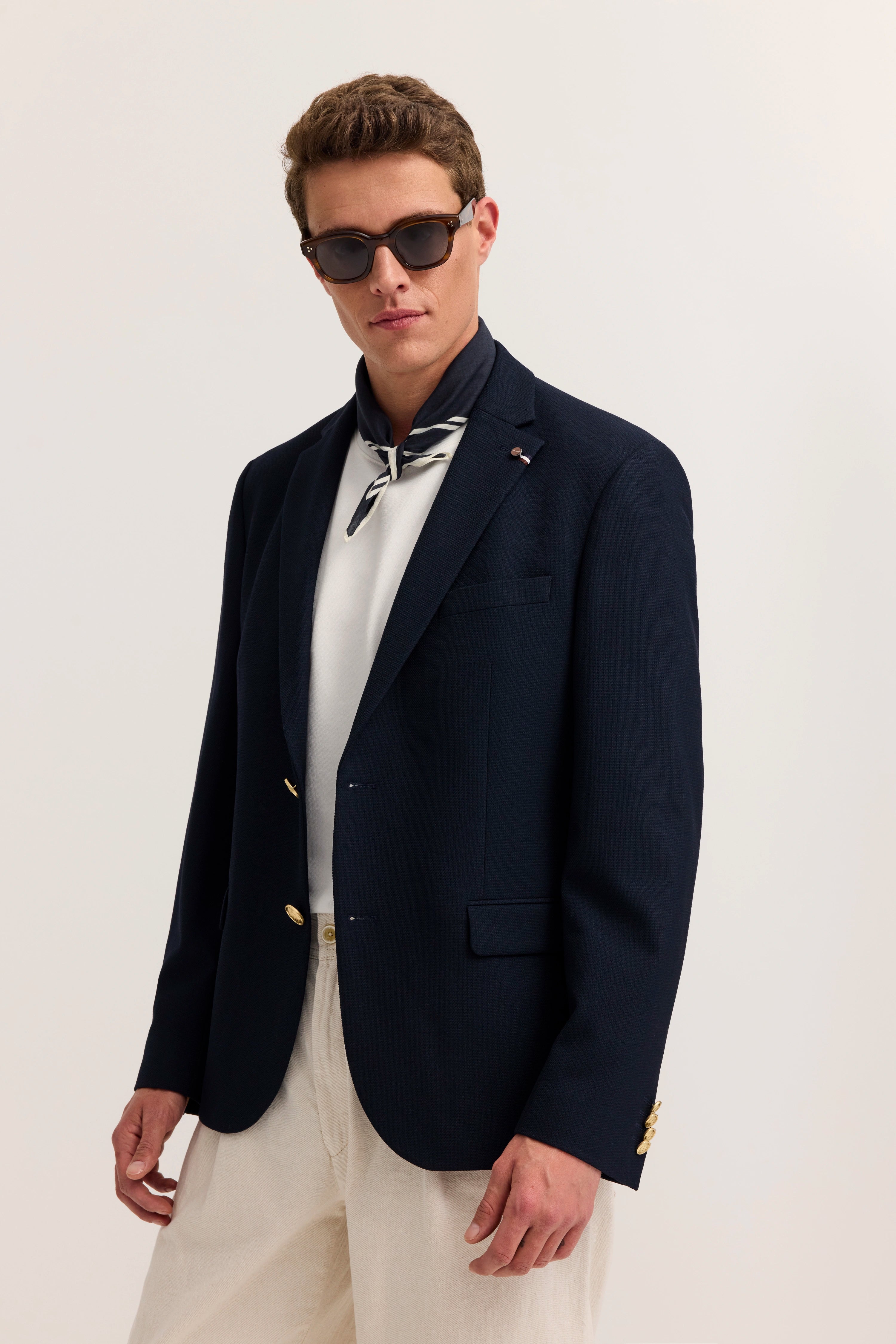 Model trägt dunkelblaues Sakko, weißes Shirt, beiges Tuch, posiert lässig mit Sonnenbrille.