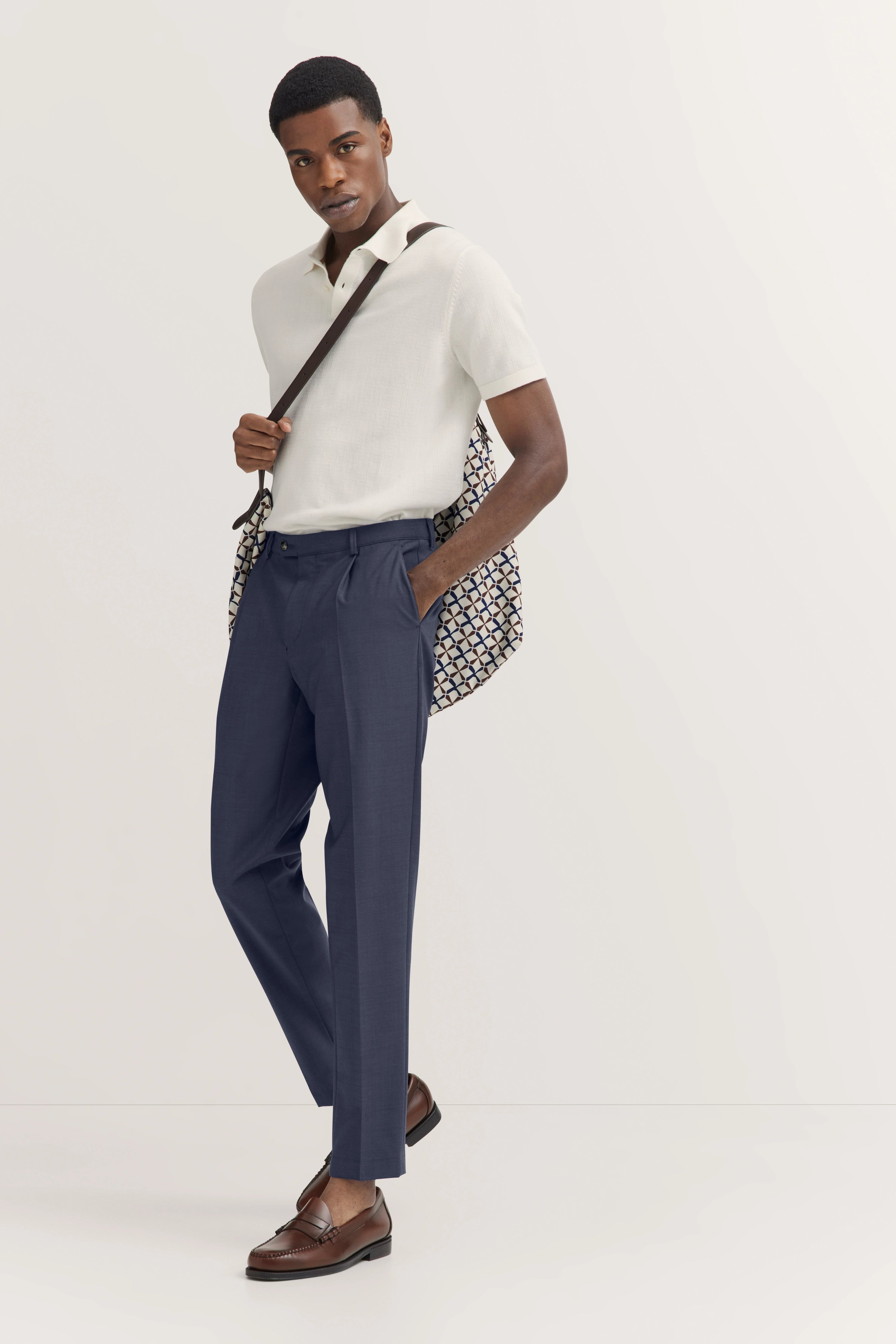Wit poloshirt, blauwe broek, bruine schoenen. Model met tas over schouder, ontspannen pose.