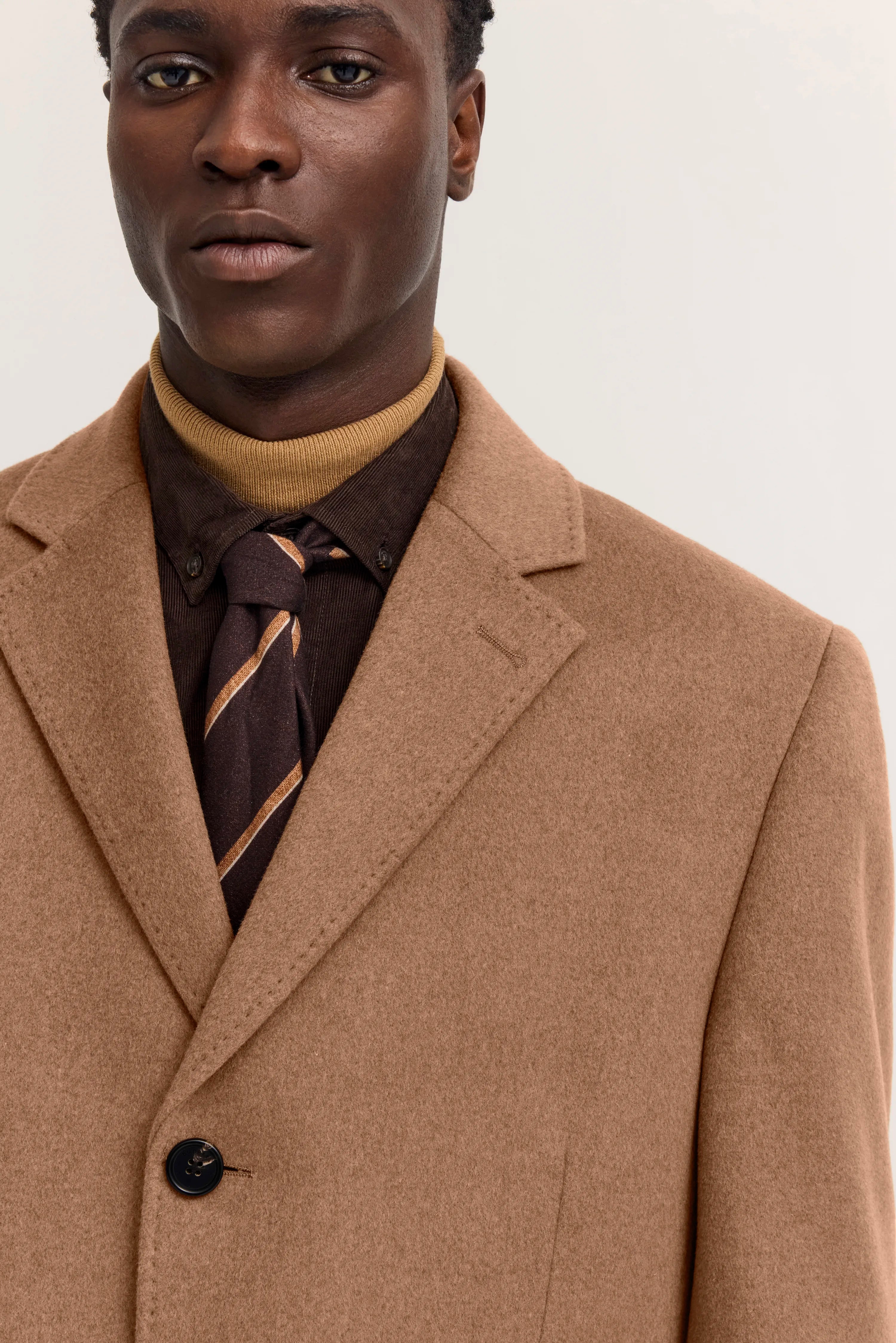 Büste eines Models in camelbraunem bugatti Mantel, braunes Hemd, senfgelber Rollkragen, gestreifte Krawatte.